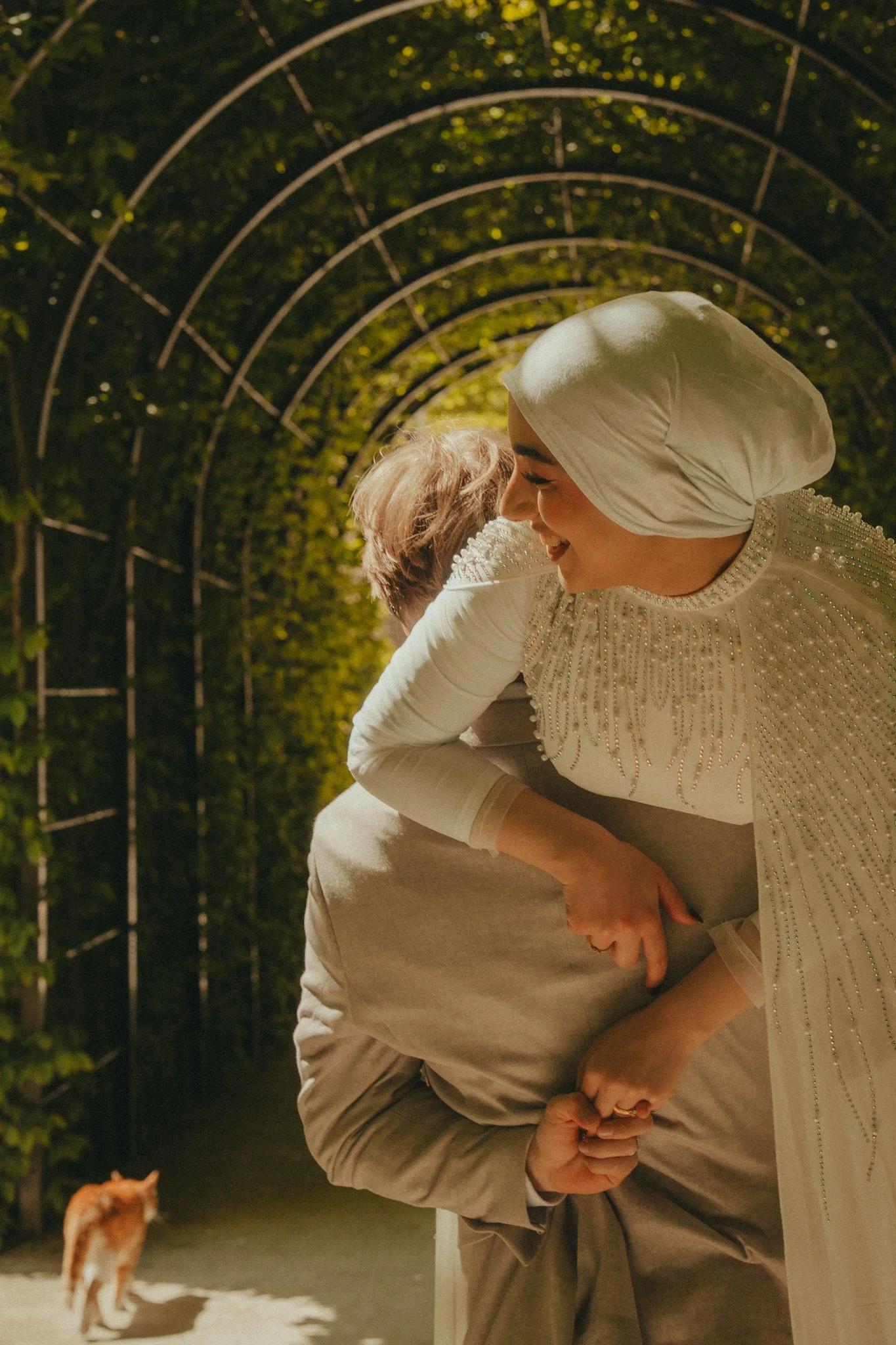 A woman dressed in a white beaded dress and white headscarf hugging a person wearing a beige jacket outside at night, with green foliage and a metal archway in the background. There is a dog in the foreground.