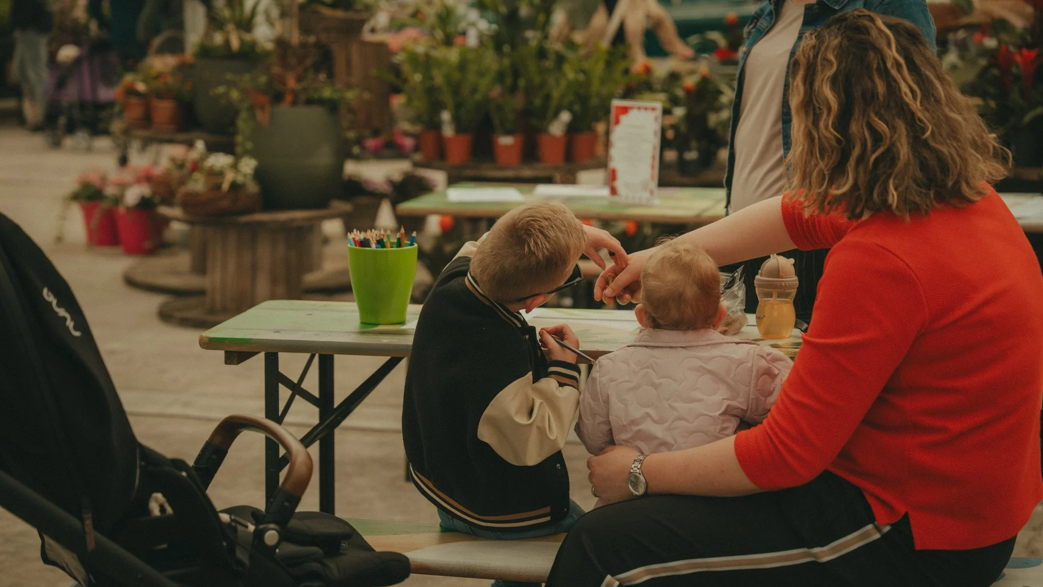 A woman in a red shirt sitting at a table with two children, one in a beige jacket and the other in a black and white striped jacket, at an outdoor setting with potted plants and flowers in the background.