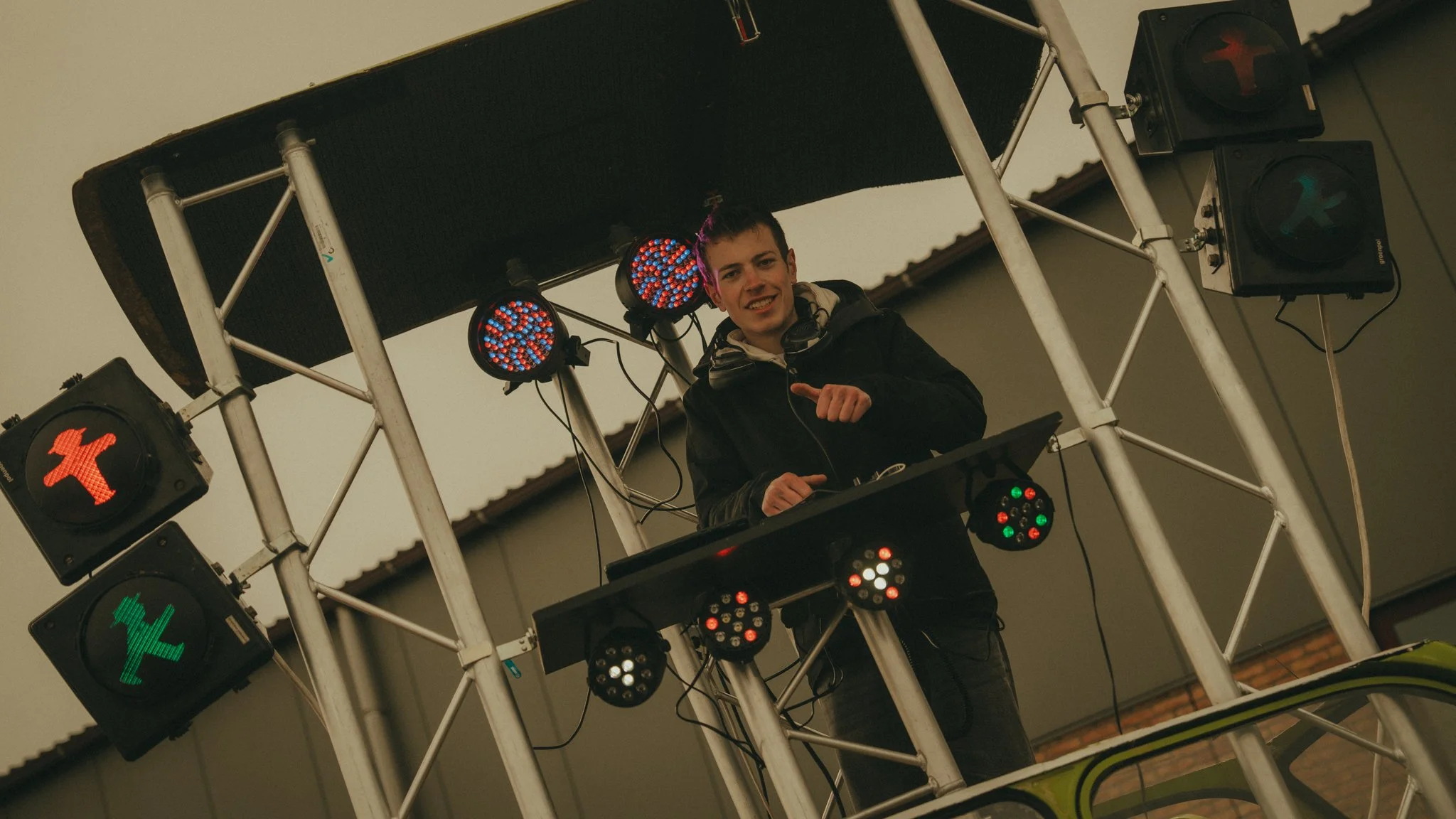 A young male DJ with headphones behind a DJ booth on a metal platform, surrounded by large colored stage lights showing green, red, and blue signals.