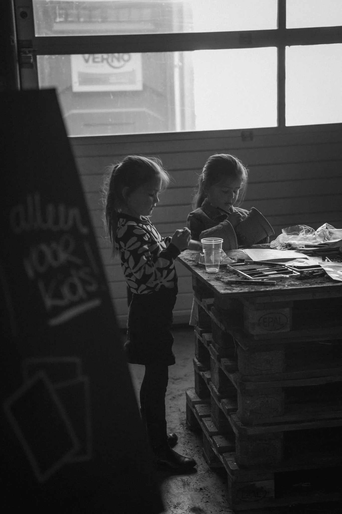 Two young girls with long hair, one with a floral jacket and the other with a vest, are standing at a worktable, engaged in writing or drawing. The scene is indoors with a large window or glass wall in the background and objects like plastic cups and