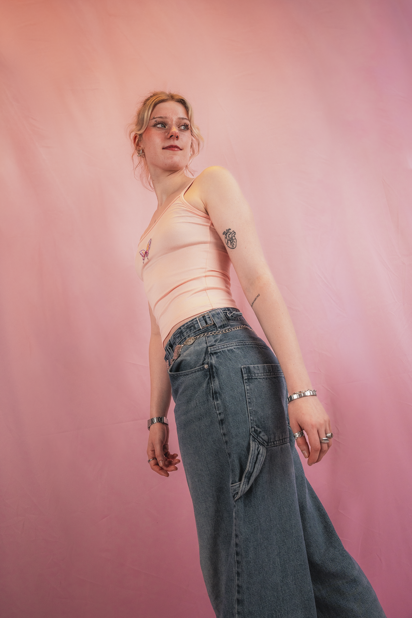 A young woman with blonde hair and fair skin posing against a pink backdrop. She is wearing a light peach tank top with a butterfly design, loose blue jeans with a chain belt, and jewelry including rings, bracelets, and earrings.