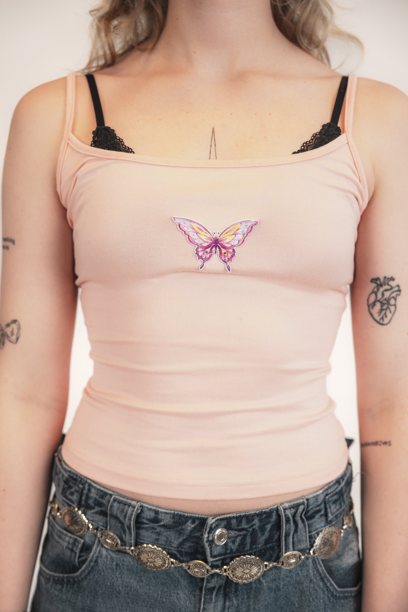 Close-up of a woman wearing a peach-colored tank top with a butterfly embroidery on the chest, black lace bra straps visible, and silver coin belt with intricate designs around her waist, with tattoos on her arms.