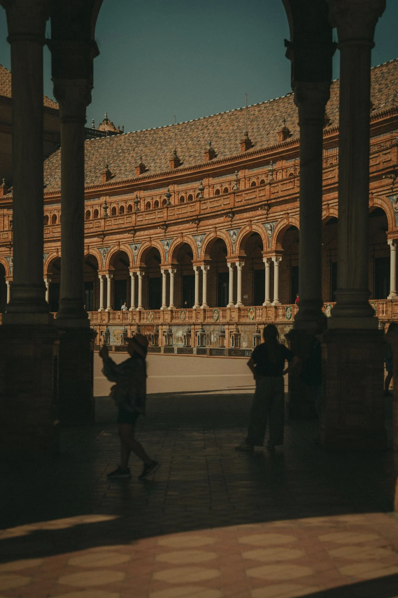 A view through arched columns showing a historic building with ornate brickwork, multiple balconies, and a tiled roof, with people walking in the courtyard.