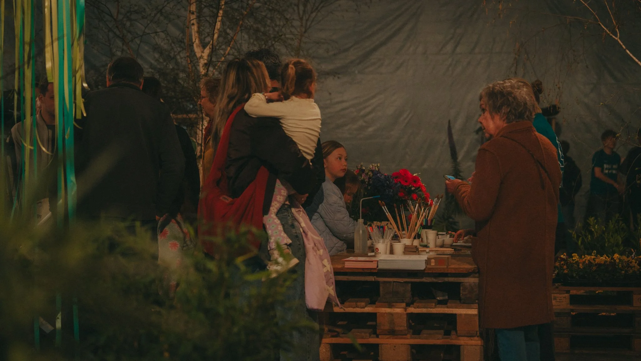 People gathered at an outdoor market or fair at night, with a table of flowers and art supplies. Some people are chatting and looking at items, with a child sitting in front of the table.