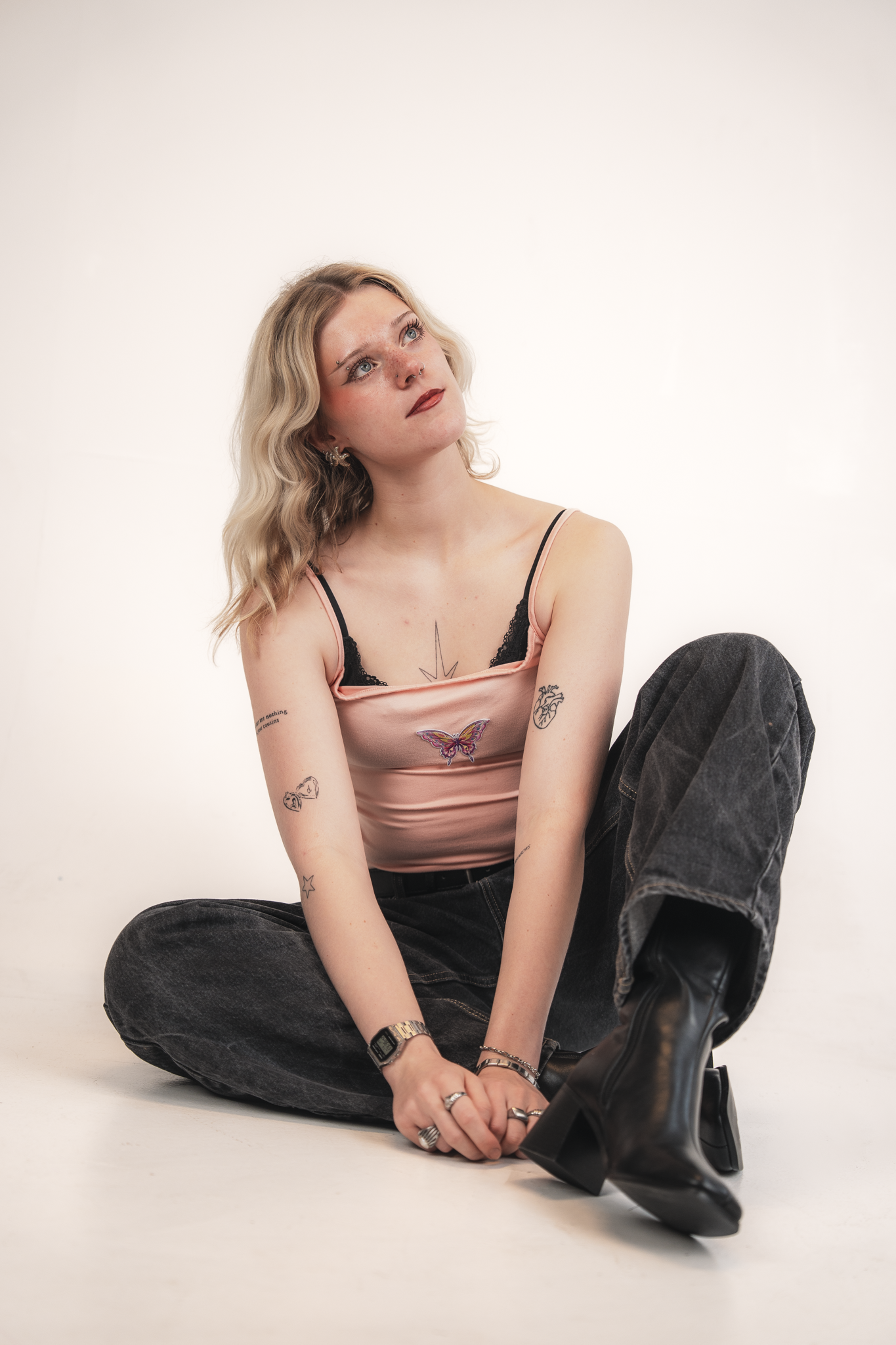 A young woman with blonde wavy hair and tattoos on her arms, sitting on the floor with her legs crossed. She wears a pink tank top with black lace, black jeans, and black boots, and looks upward with a thoughtful expression.