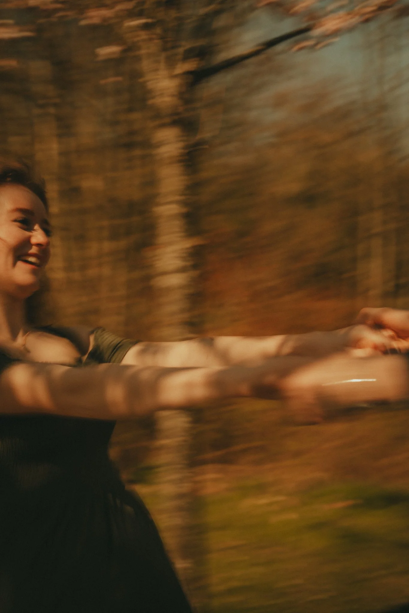 A woman smiling and extending her arm, possibly taking a selfie or reaching out, with a blurred brown background that suggests motion.