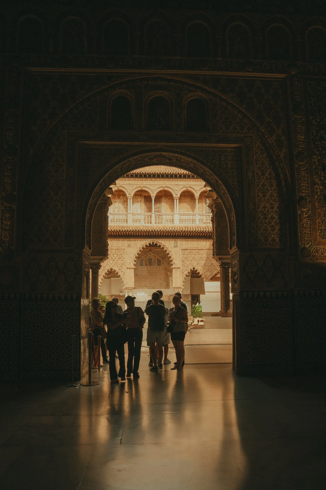 People standing and talking inside a historical building with intricate arches and carvings, illuminated by sunlight coming through the openings.