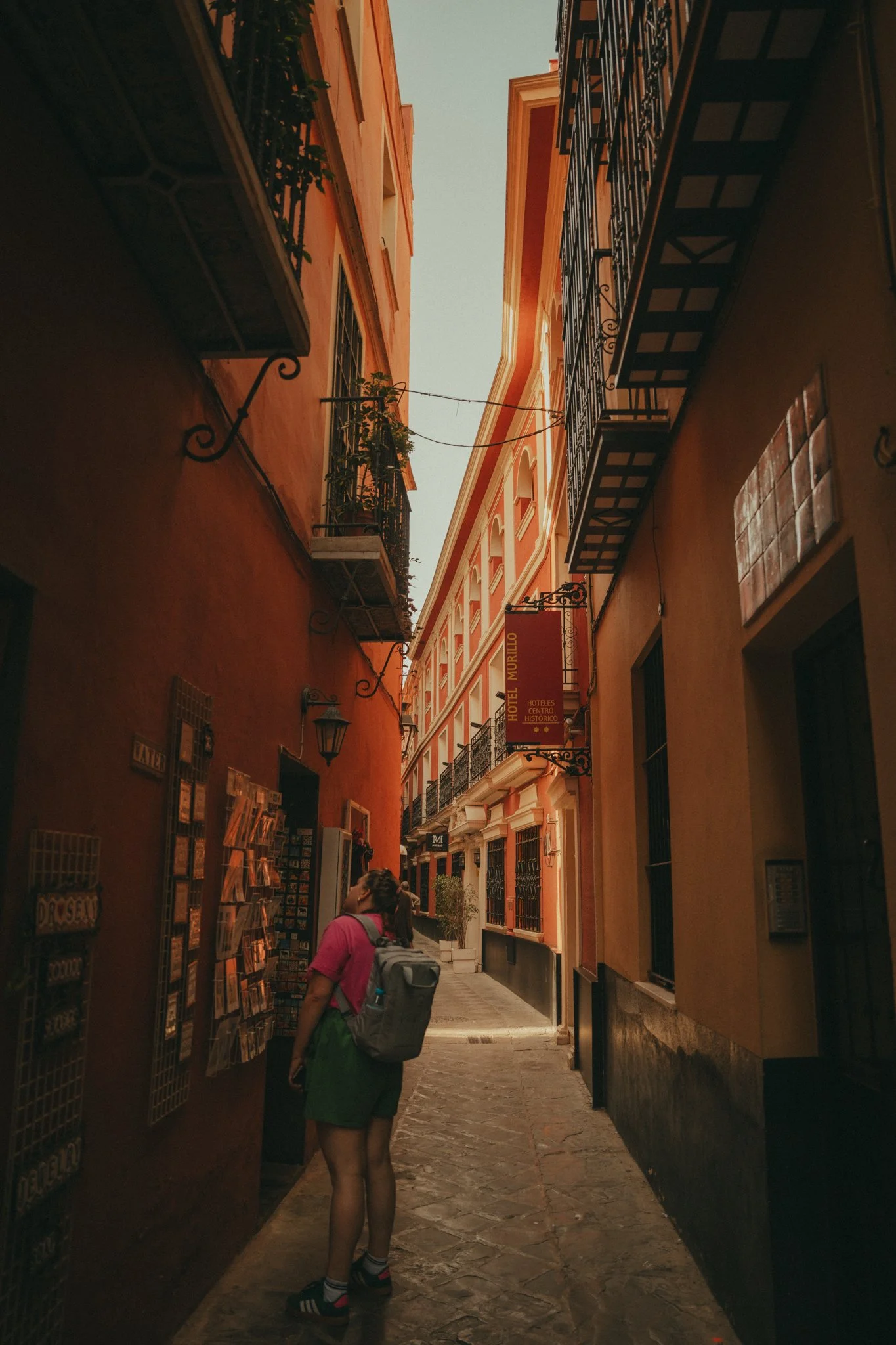 A woman with a backpack standing in a narrow cobblestone street, looking at a display rack outdoors, surrounded by colorful tall buildings with balconies and wrought-iron railings.