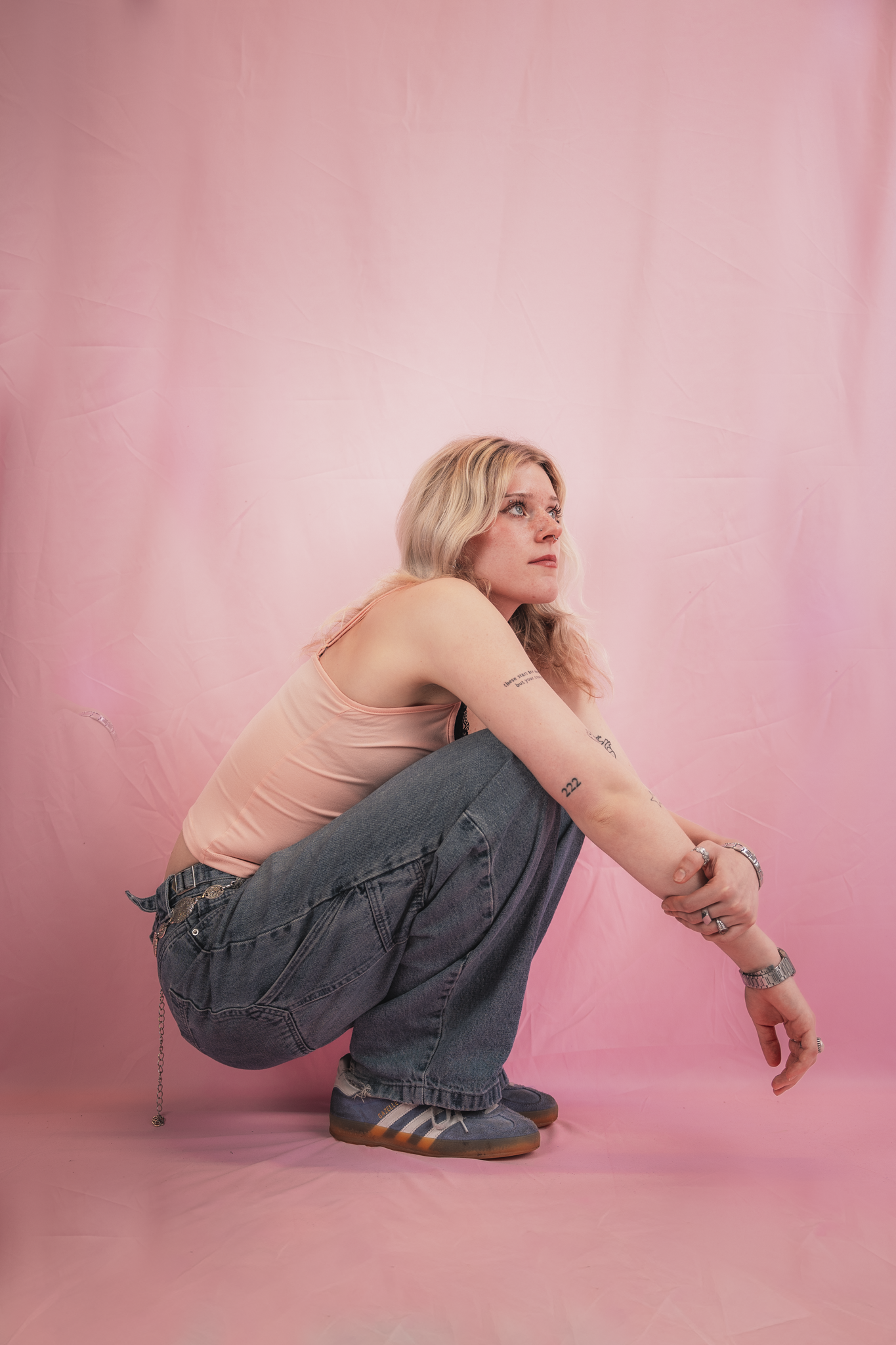 A young woman with long, wavy blonde hair, wearing a light pink tank top, blue jeans, and sneakers, is crouching against a pink backdrop. She has tattoos on her arms and is wearing jewelry and a watch, looking thoughtfully to her right.