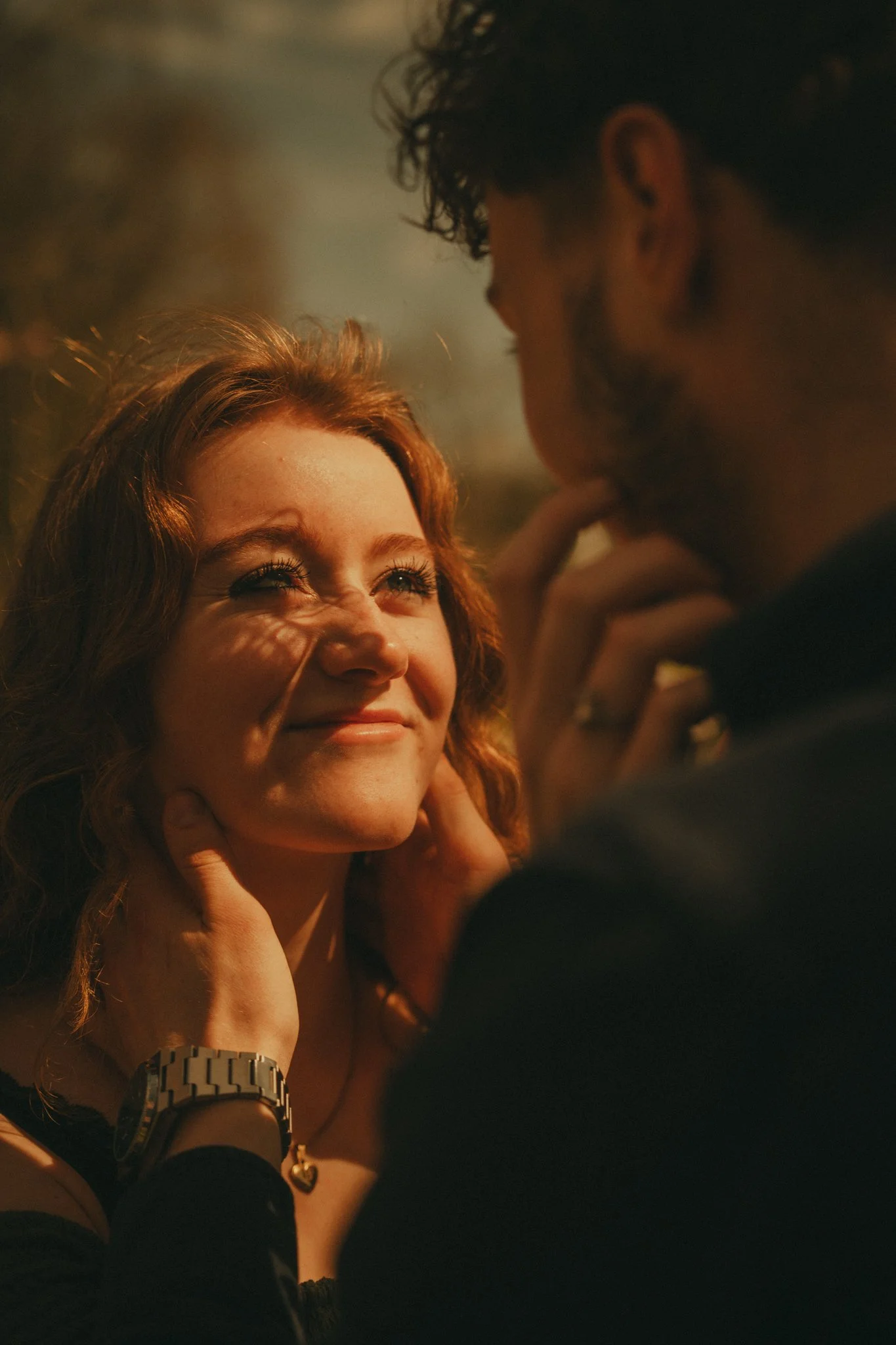 A woman with red hair smiling while a man with dark curly hair and beard stands close to her, with his hand near his mouth, in a dimly lit setting.