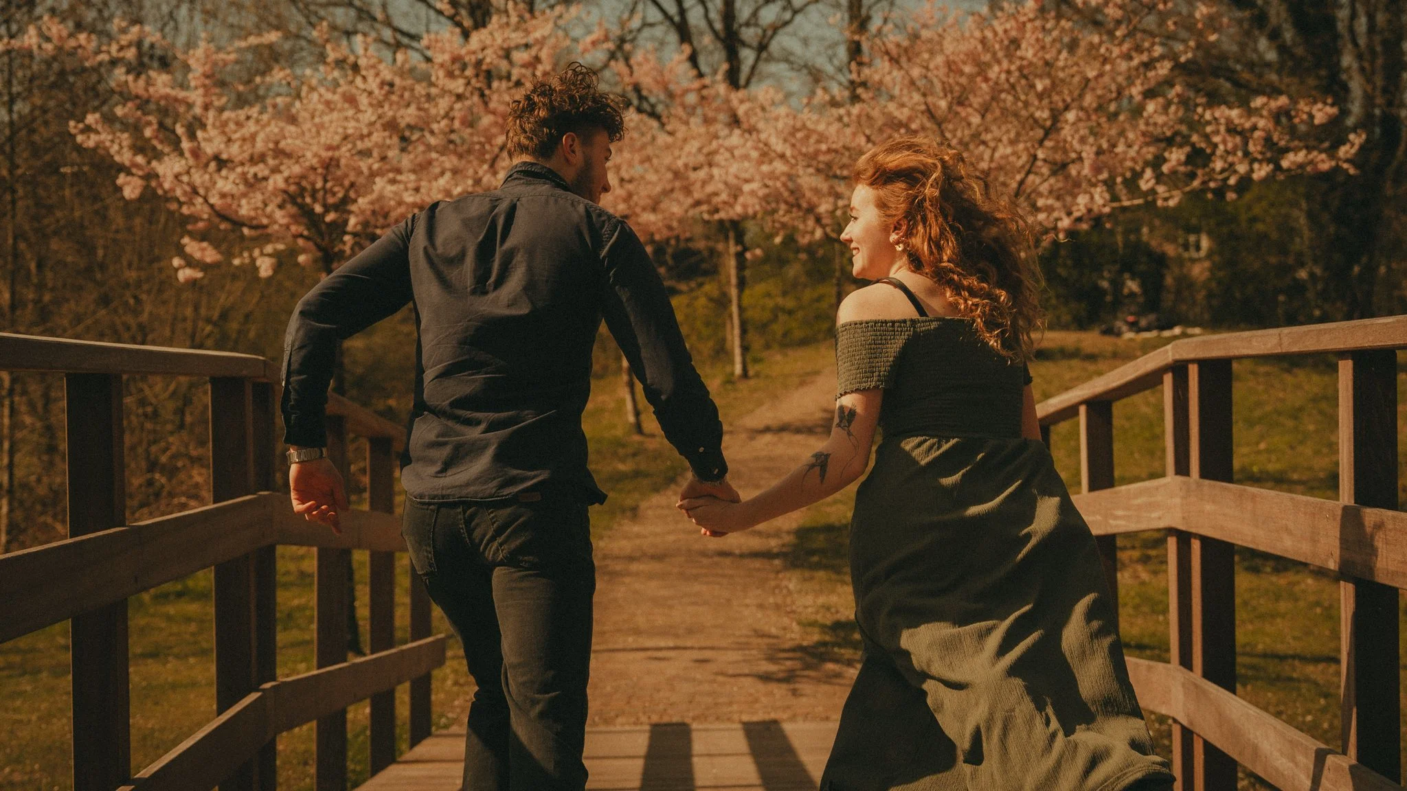 A young couple holding hands and walking together on a wooden bridge, surrounded by blooming cherry blossom trees.