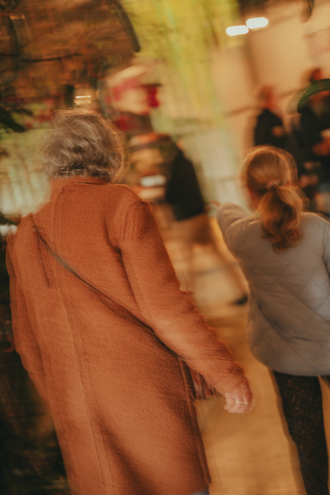 A person in an orange coat is holding hands with someone else, in an indoor setting with warm lighting and other people in the background.