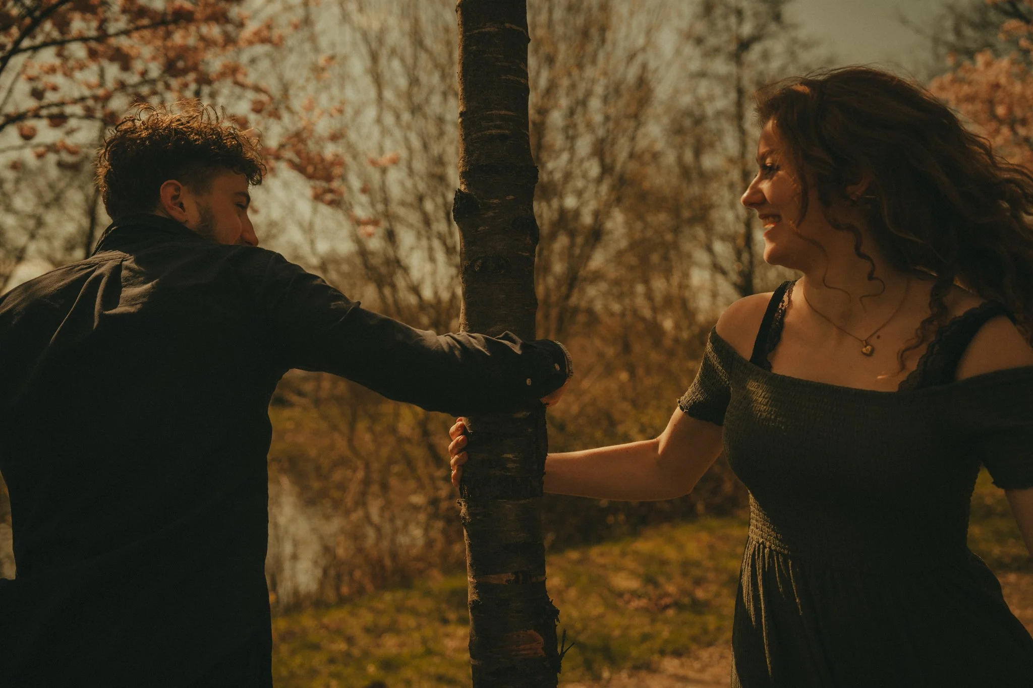 A man and a woman smiling and holding hands around a tree outdoors in autumn.