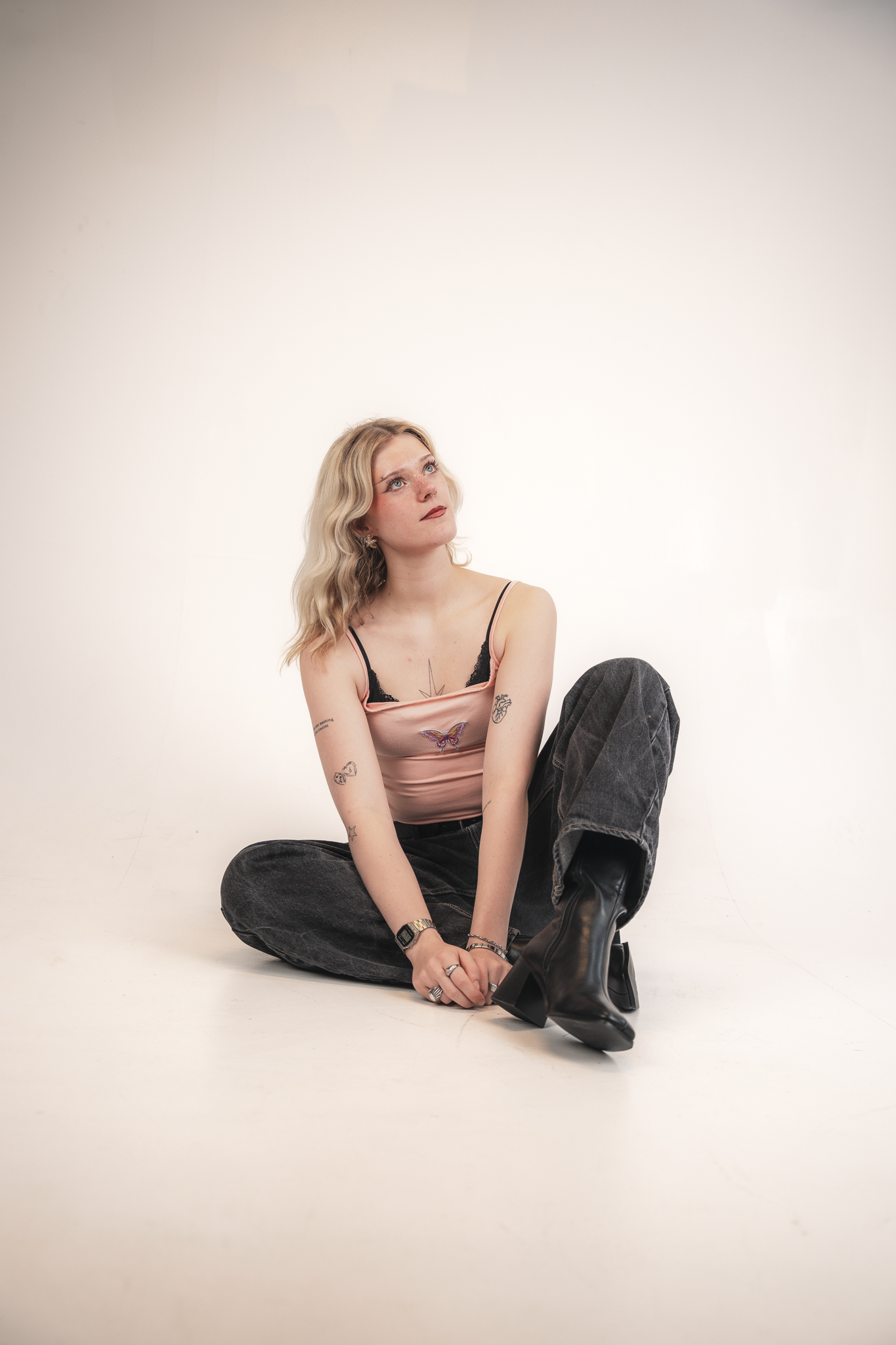 A young woman with blonde wavy hair sits on the floor against a plain white background, looking upwards. She is wearing a light pink spaghetti strap top with black lace detail, dark baggy jeans, and black heeled boots. She has various tattoos on her 