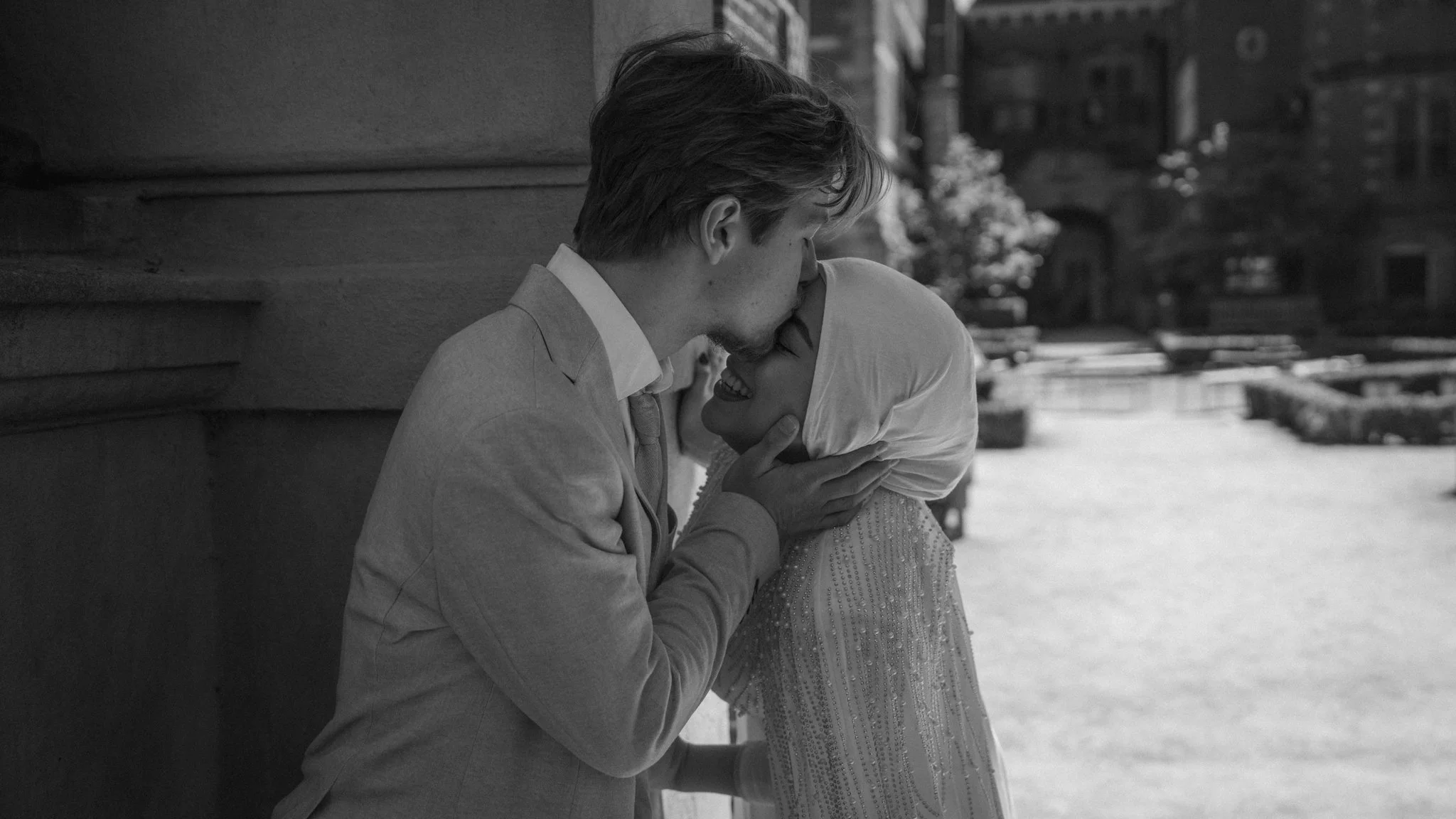 A black and white photo of a couple sharing a kiss outside on a city street, leaning against a building with blurred city buildings in the background.