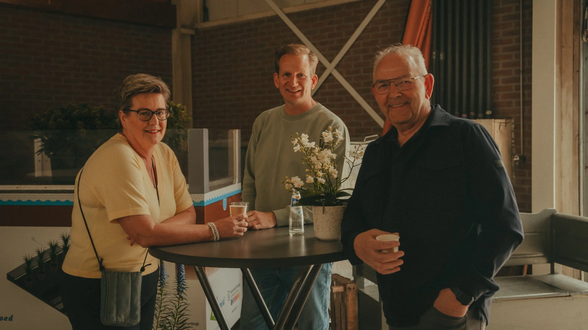 Three adults standing together at a social gathering, smiling, with a woman in a yellow top, a man with blond hair in a gray sweater, and an older man in black holding a coffee cup, near a plant with white flowers.
