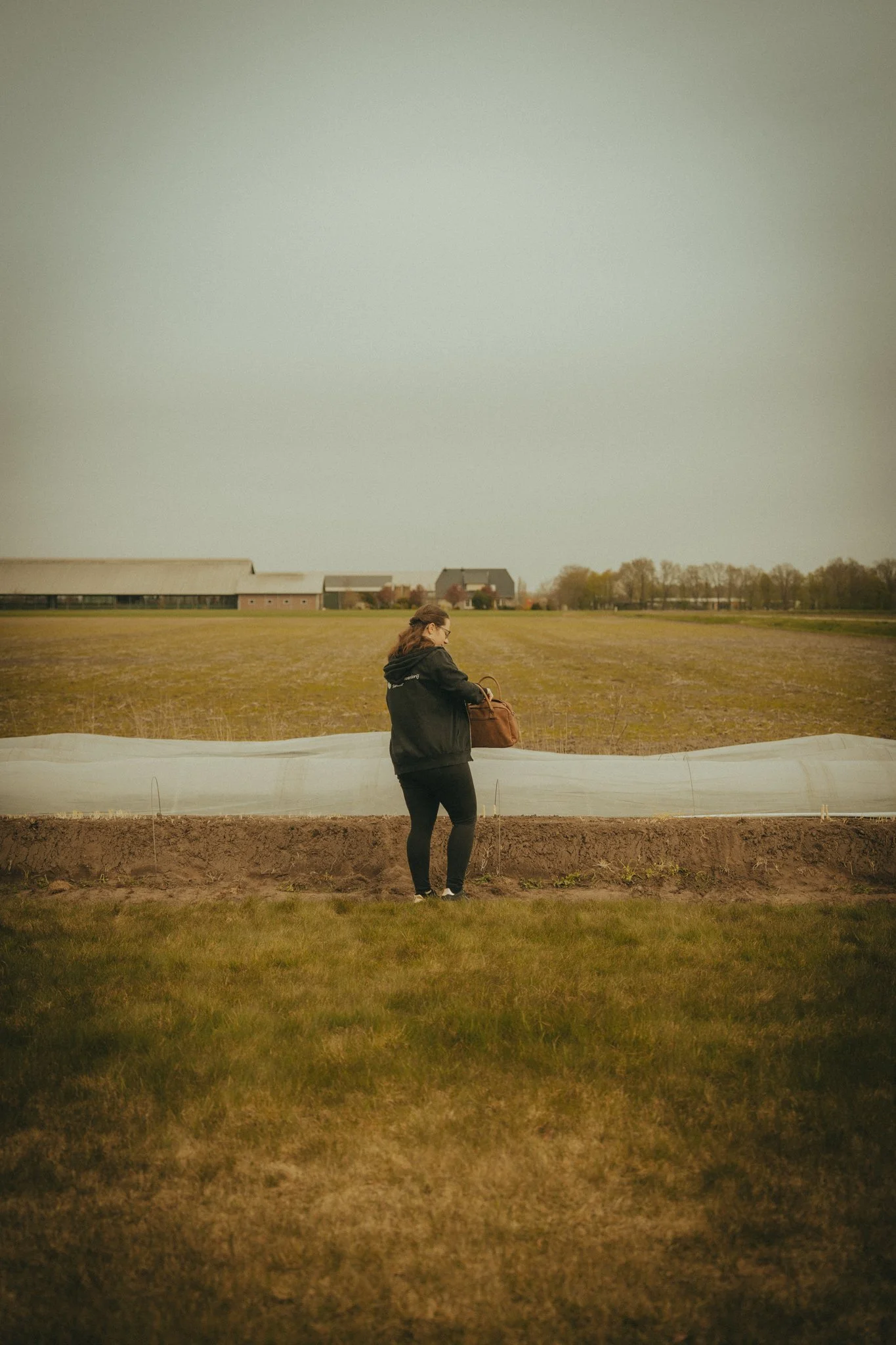 A woman near a farm field holds a brown bag, with farm buildings and trees in the background under an overcast sky.