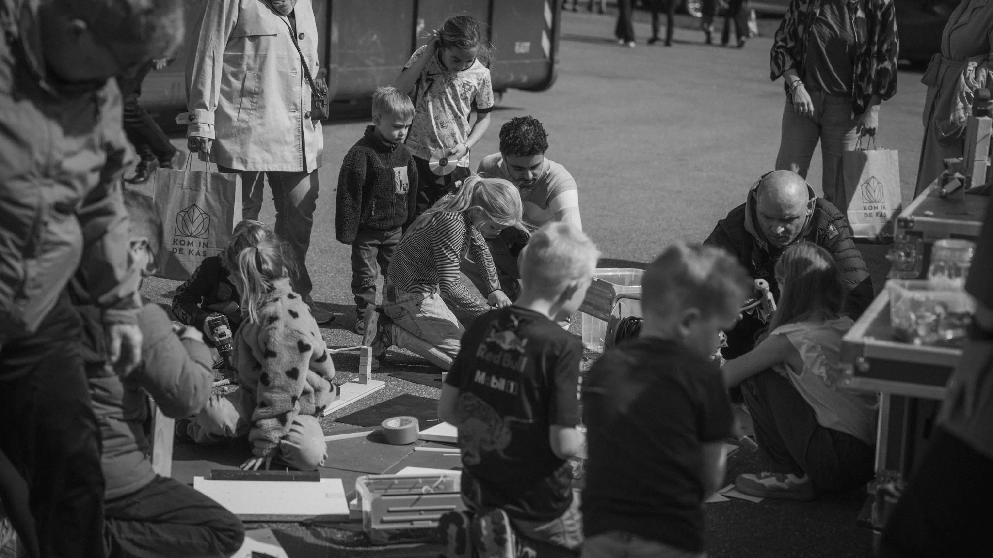A group of children and adults engaging in an outdoor activity at a market or fair, with some children sitting on the ground and others standing around, surrounded by tables and bags.