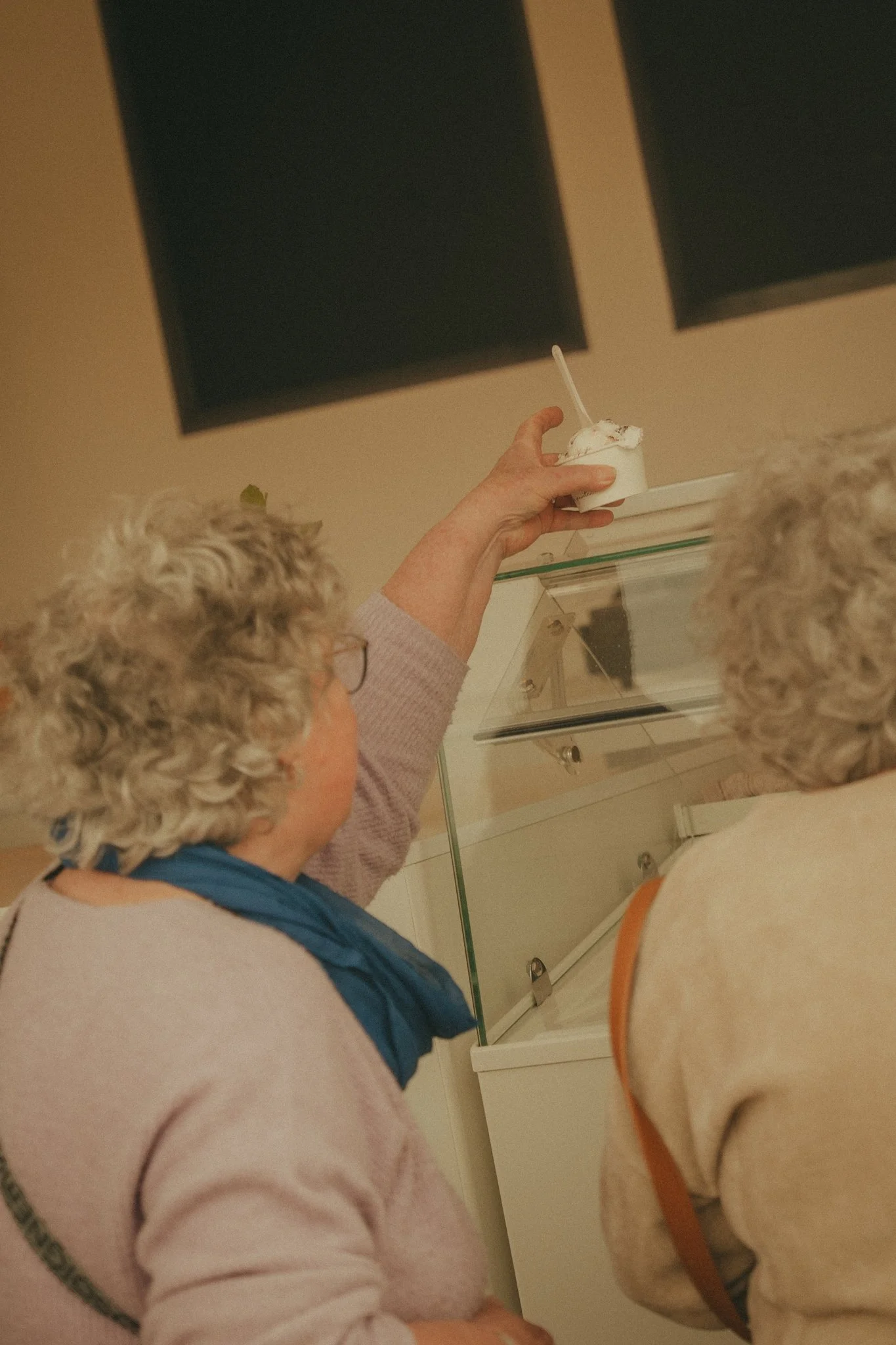 An elderly person with curly gray hair is seen at a store or cafe, holding a small cup of ice cream with a spoon. There is another person in the background and a glass display case nearby.