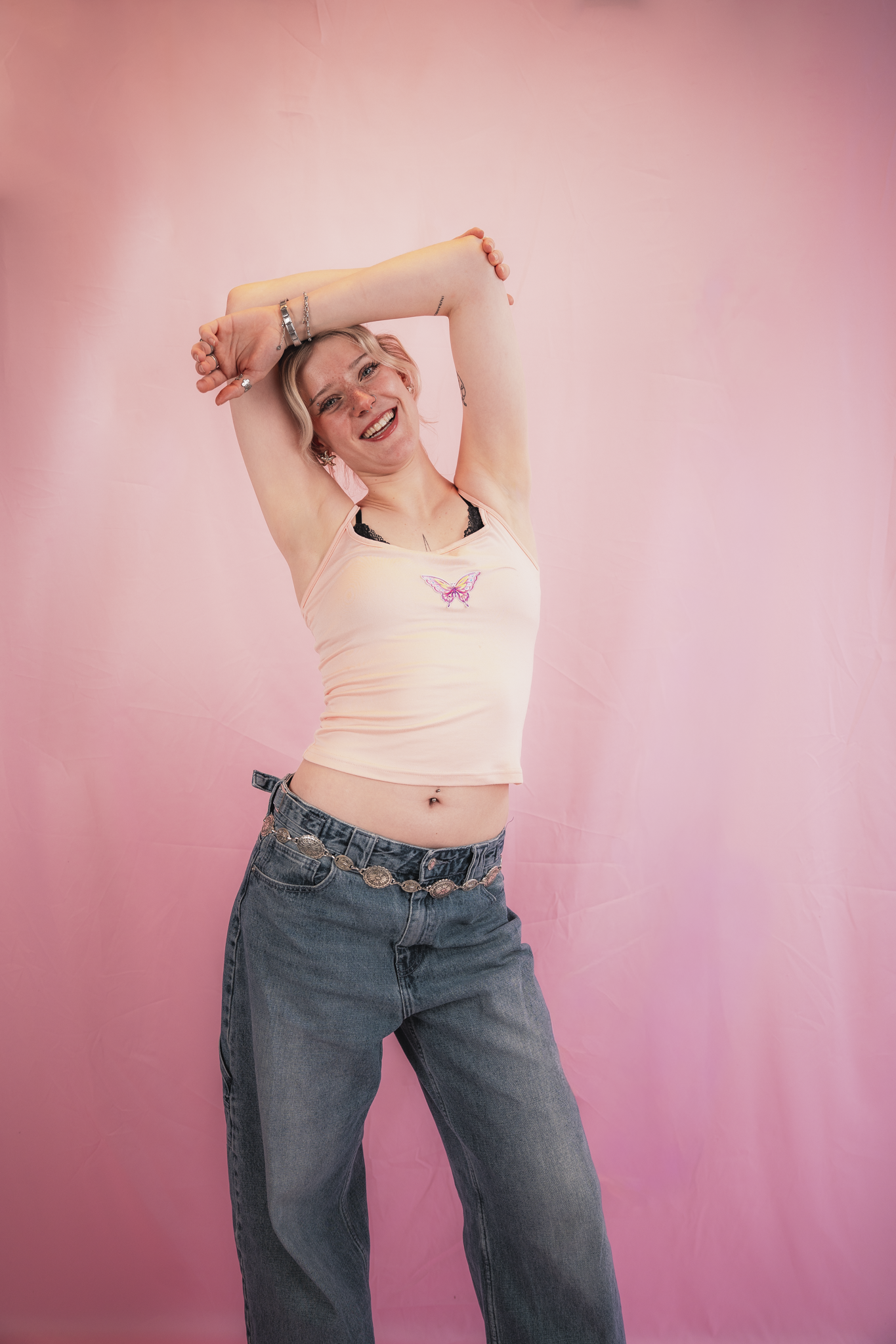 A young woman with blonde hair smiles while standing against a pink background. She is wearing a light pink tank top with a butterfly design, blue jeans, and silver jewelry, and has her arms raised above her head in a playful pose.