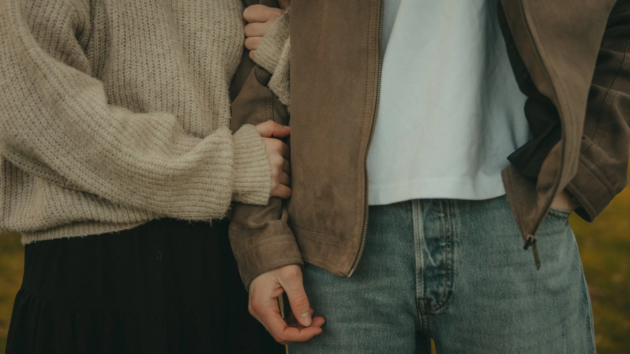 Two people holding hands, one wearing a beige sweater and the other wearing denim jeans and a brown jacket.