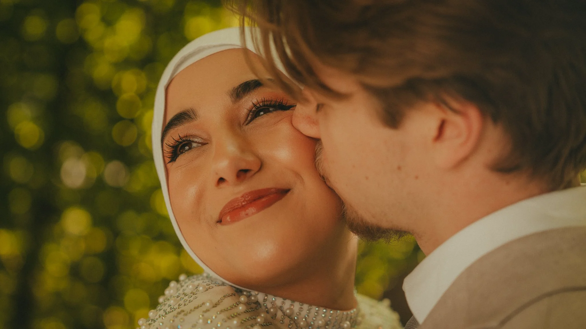 A woman wearing a white headscarf and pearl jewelry looking at a man with her reflection visible in a mirror, with a blurred golden bokeh background.