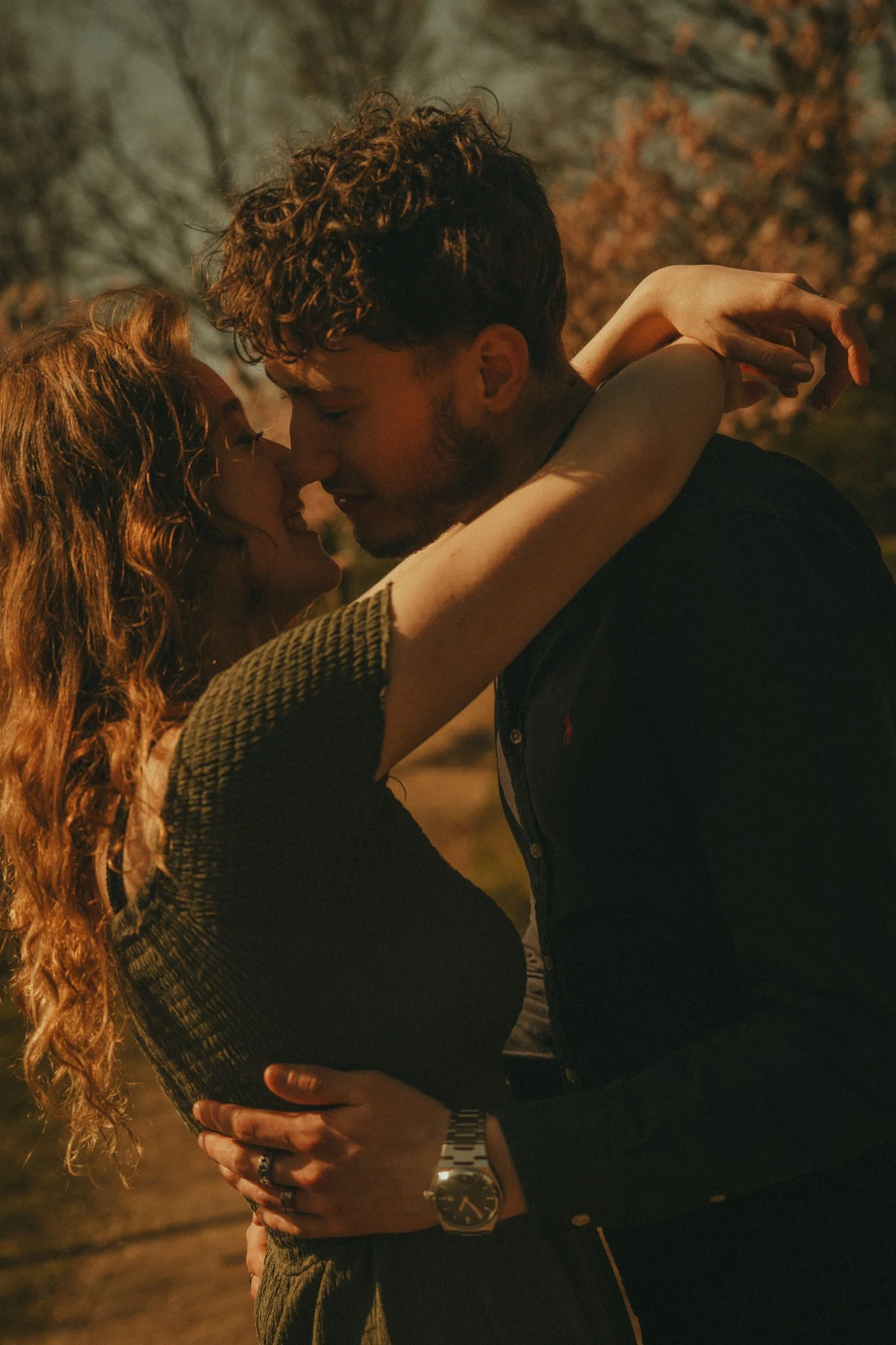 A young couple is embracing and about to kiss outdoors during sunset, with trees in the background.