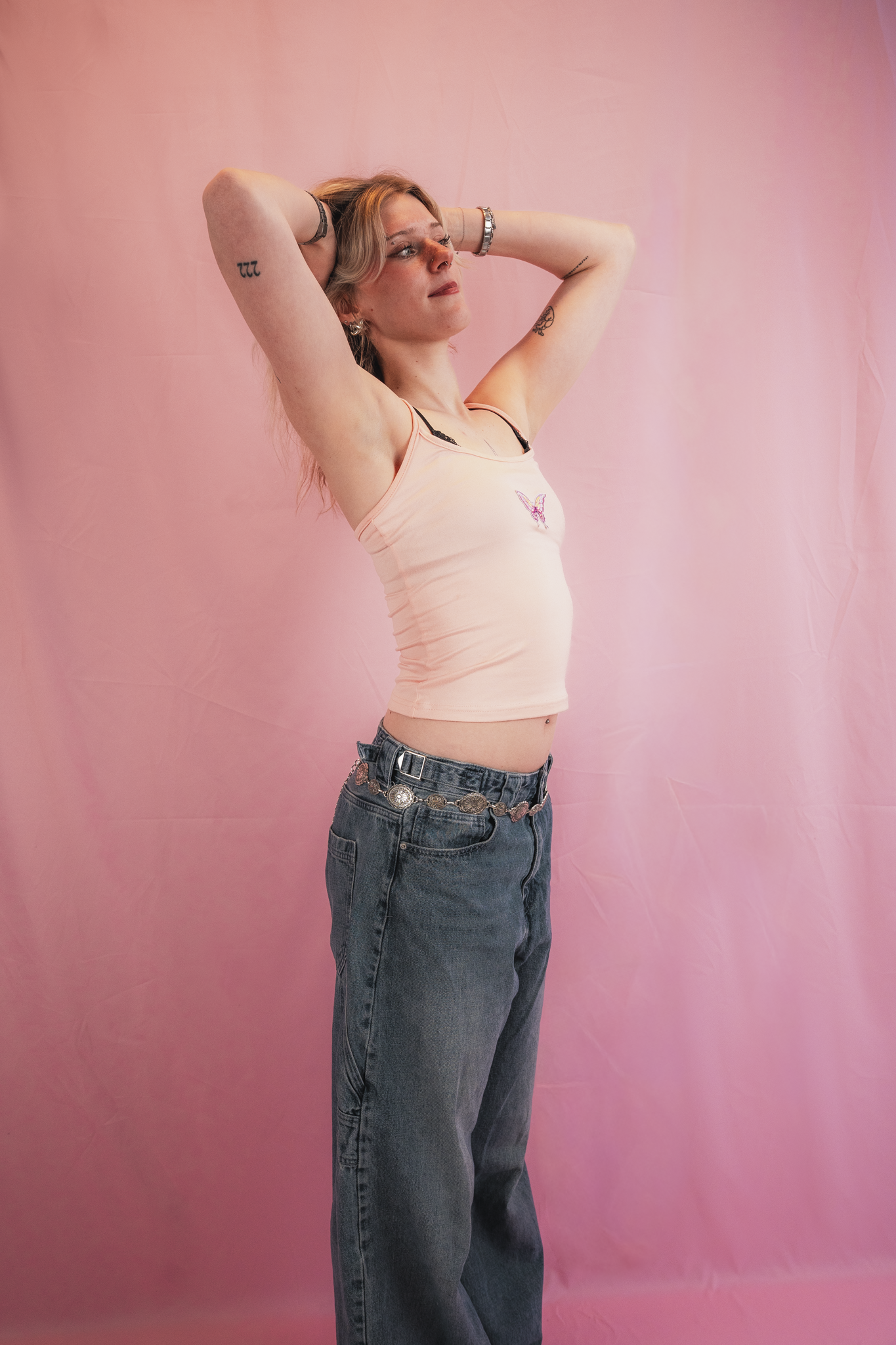 Young woman with blonde hair posing against a pink background with her arms raised behind her head, wearing a light pink tank top with a butterfly graphic and dark jeans, accessorized with jewelry and tattoos on her arms.
