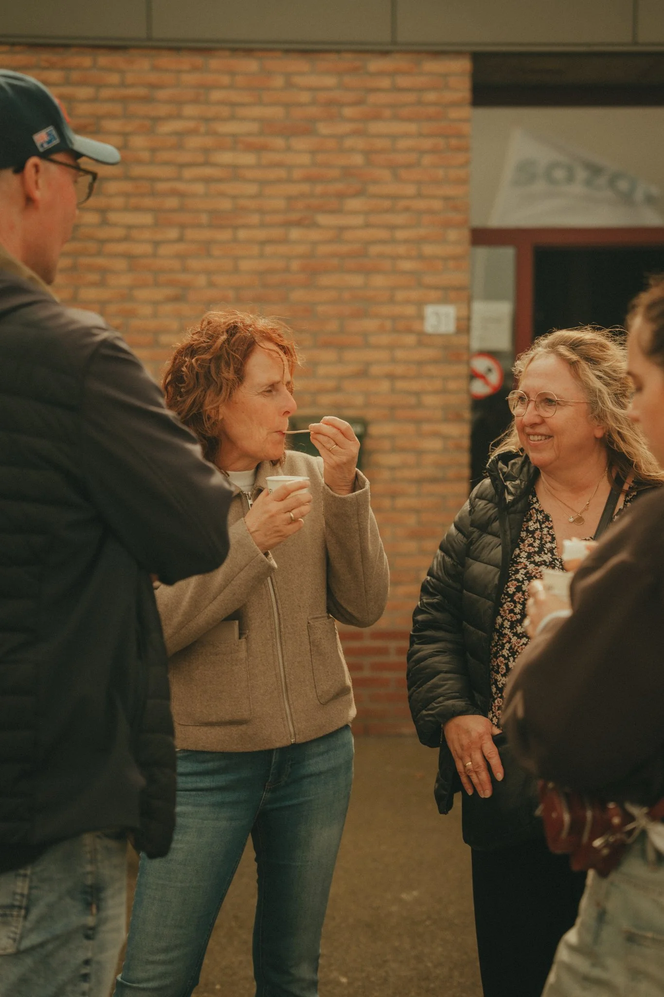 Group of people socializing indoors in front of brick wall, some smiling, one woman drinking, others partly visible.