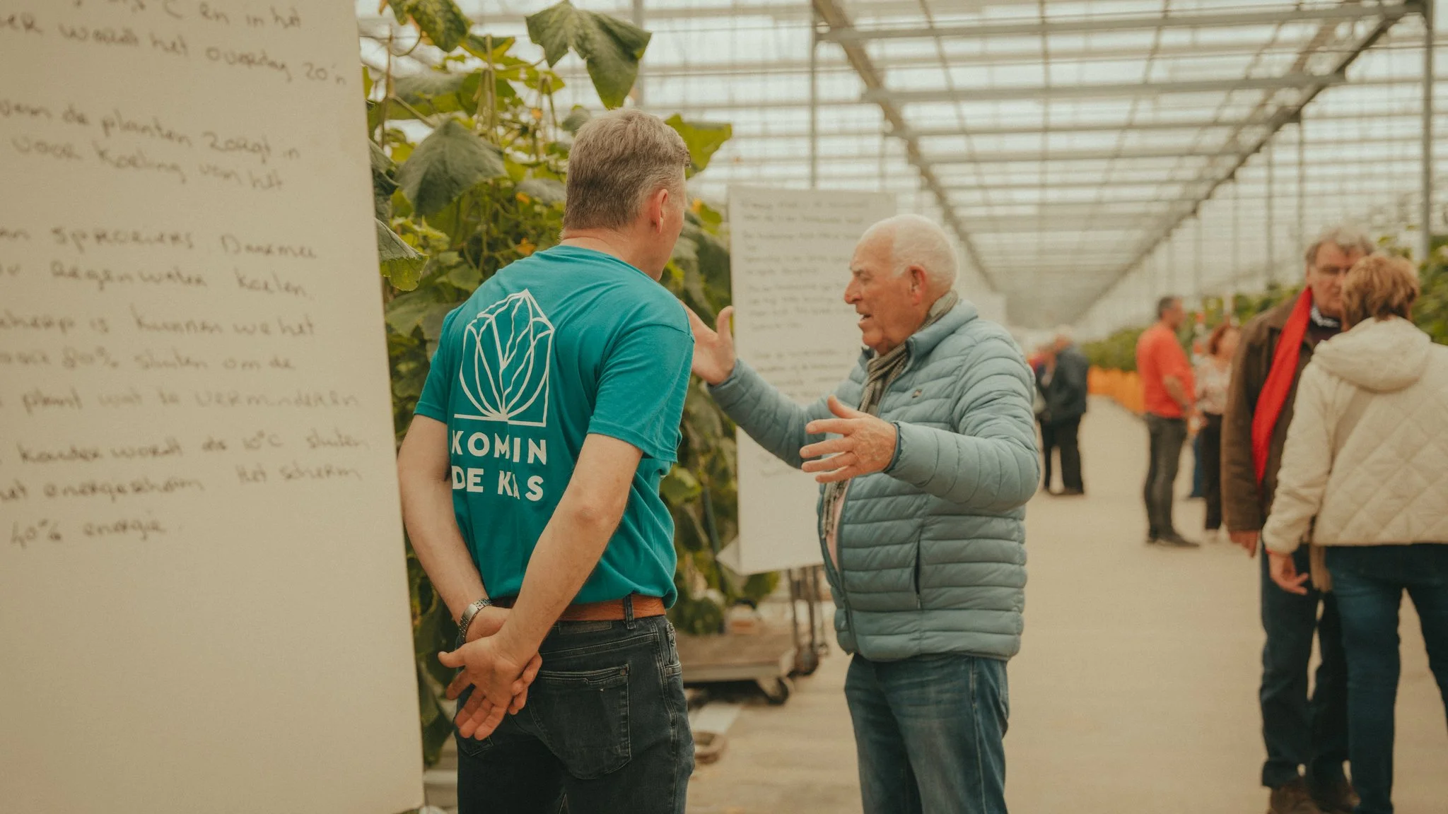 An elderly man and a middle-aged man are having a conversation inside a greenhouse or an indoor farm. The elderly man is gesturing with his hands as he talks, while the younger man listens. There are other people in the background, and informational 
