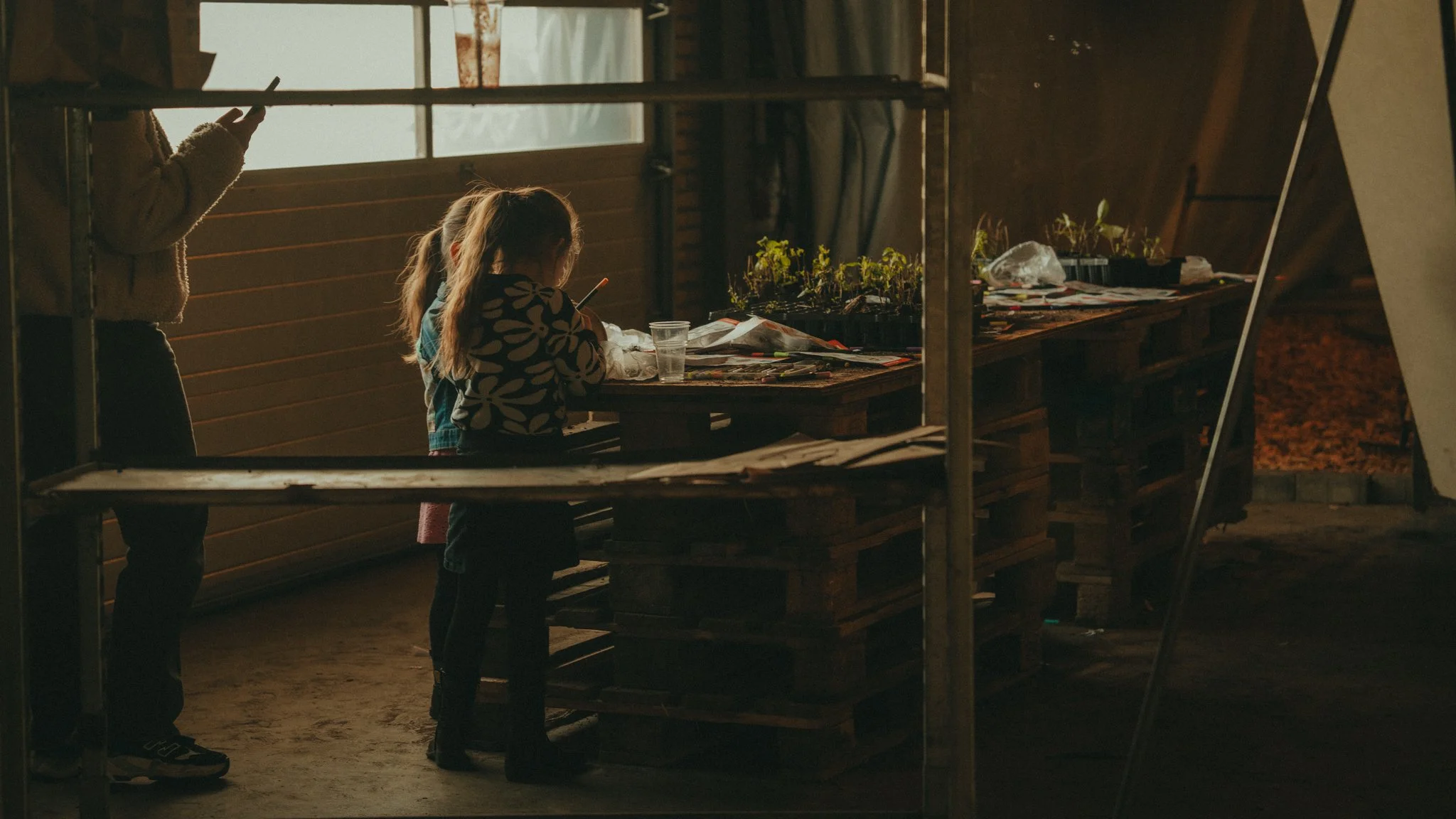 Two children, a girl with a ponytail and a boy with a sleeve, are inside a rustic wooden greenhouse or shed, tending to small plants on a table with gardening supplies. The scene is lit by natural light coming through the window.