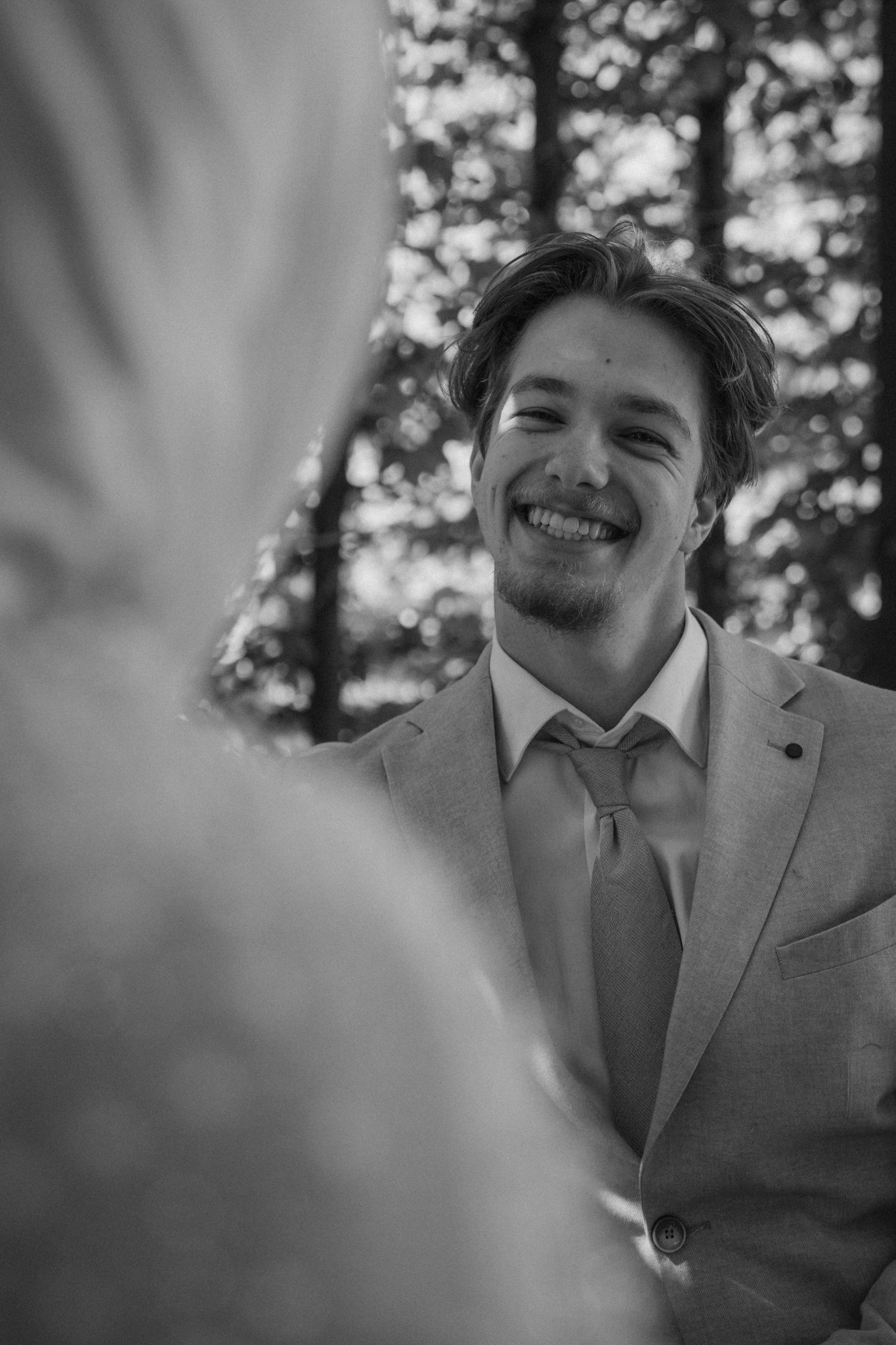 A young man in a light-colored suit and tie smiling outdoors, with trees blurred in the background, black and white photograph.