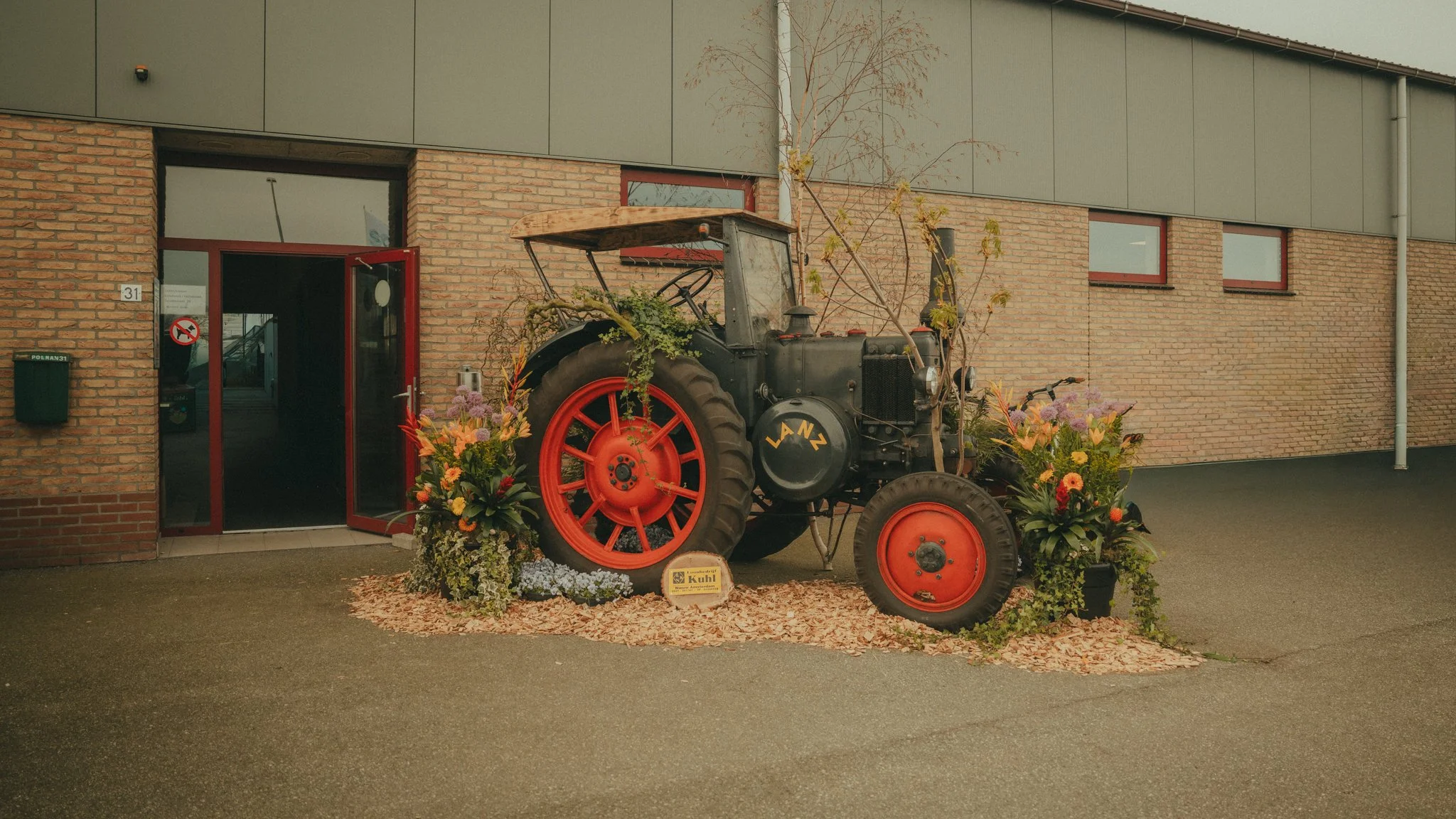 An antique tractor decorated with flowers is placed outside a brick building with a modern upper facade. The tractor has red wheels and is surrounded by flower arrangements and a small sign in the front.