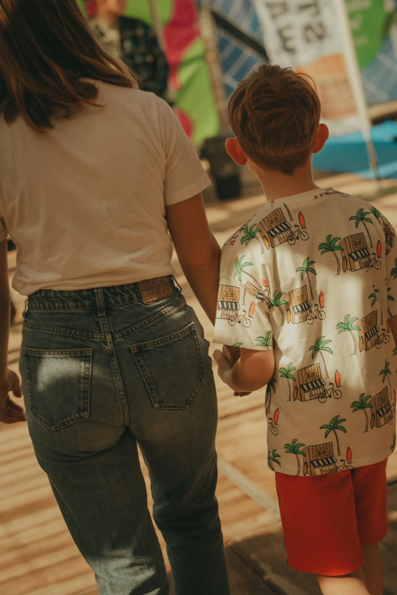 Two children walking together indoors, with a woman nearby. The girl has shoulder-length brown hair, a white T-shirt, and denim jeans. The boy has short hair, a printed T-shirt with tropical scenes, and red shorts.