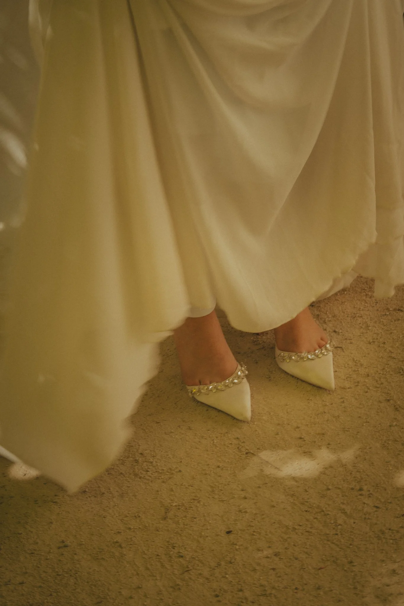 Close-up of a woman wearing a yellow satin dress with pearl and rhinestone embellished white shoes, standing on a carpeted floor.