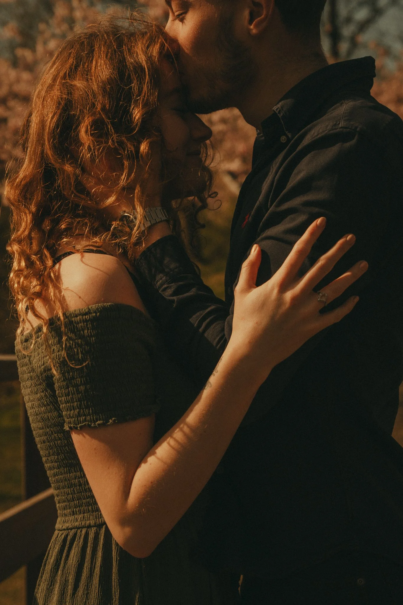 A man and a woman are close together, with the man gently kissing the woman's forehead. The woman has curly red hair and is wearing a dark off-shoulder top, while the man has short dark hair and is wearing a black shirt. The scene appears to be outdo