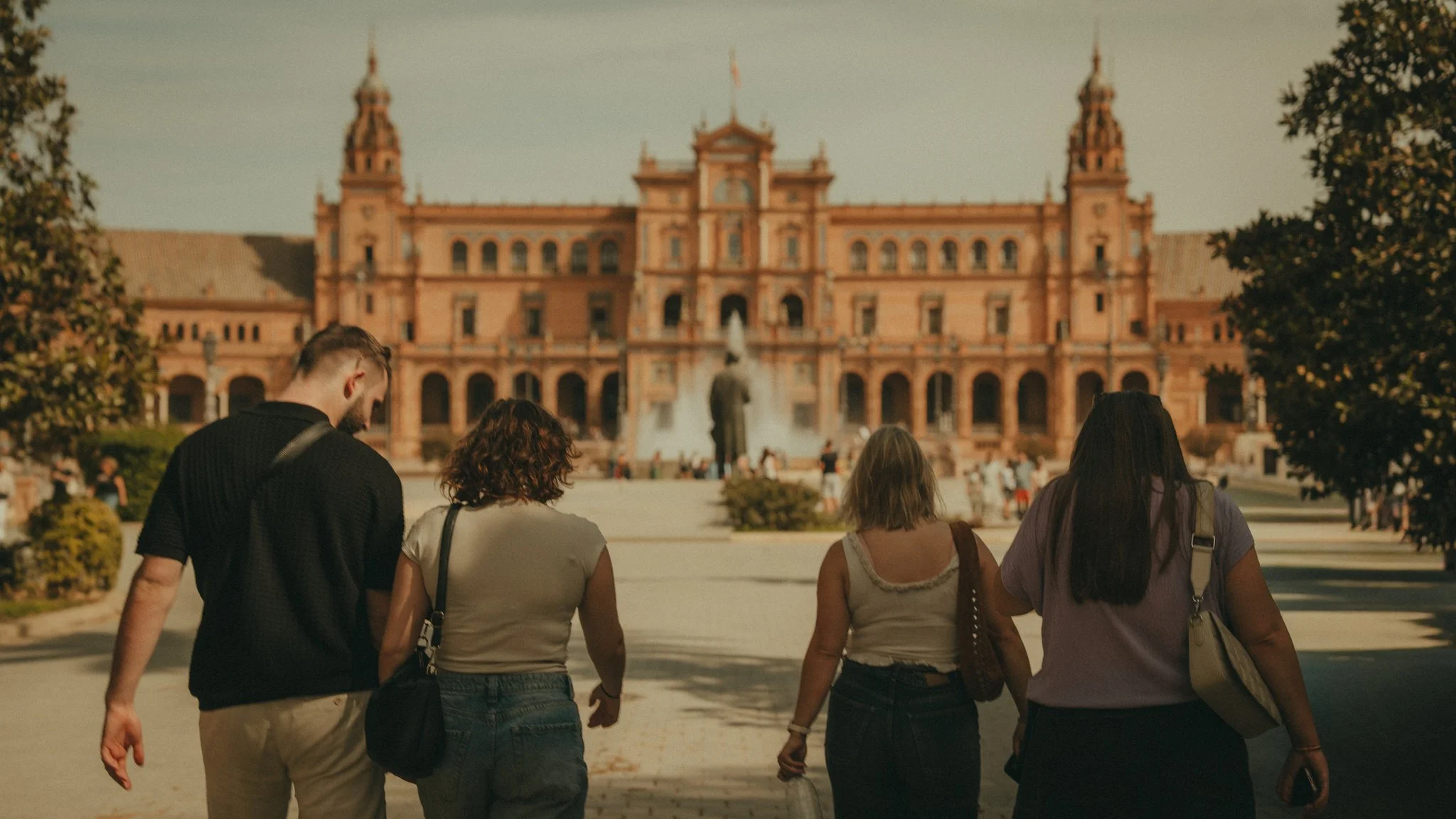 Group of four people walking towards a historic pink building with towers and a fountain in front.