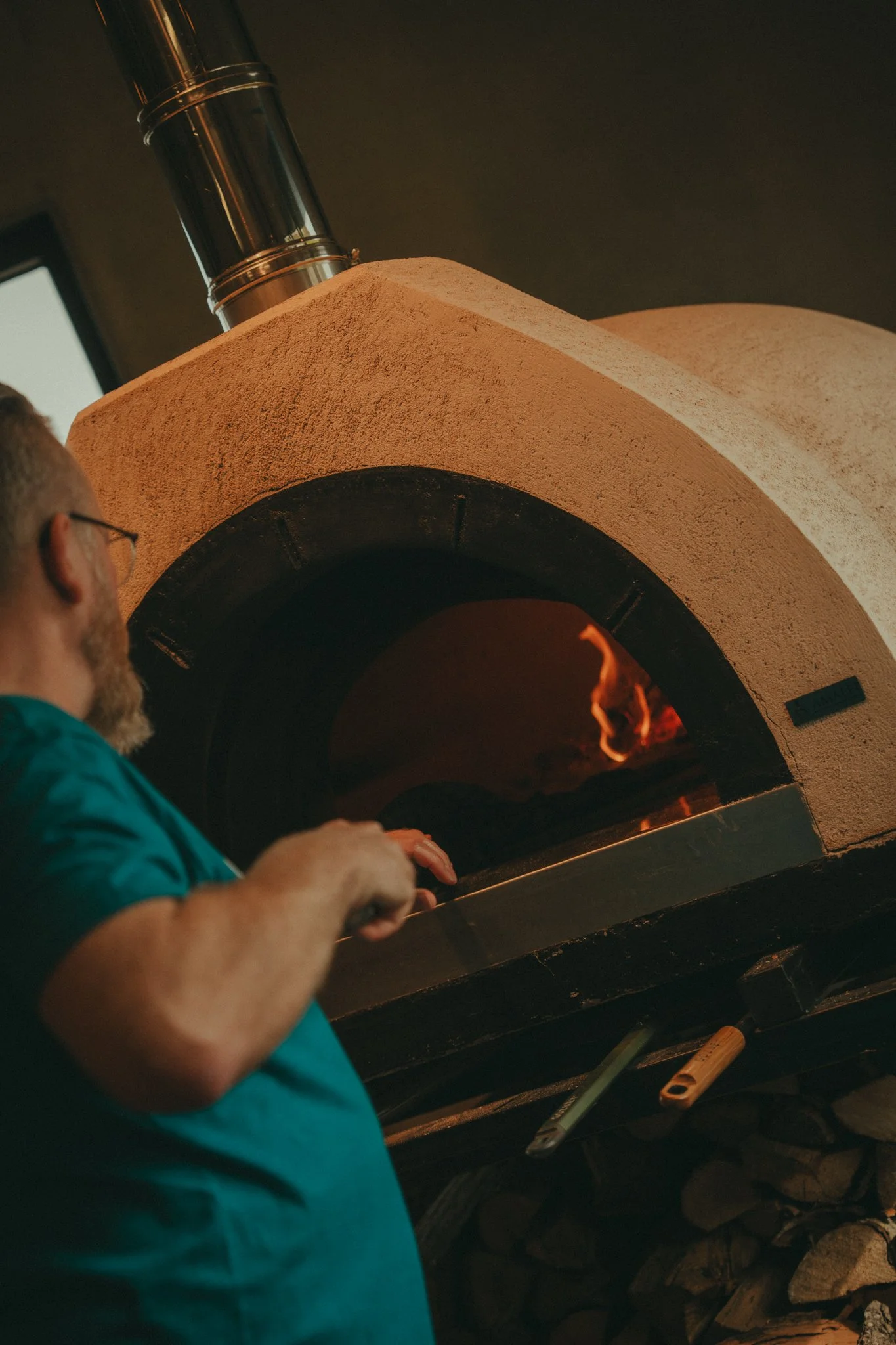 A person wearing glasses and a teal shirt is tending a fire inside a stone oven or kiln.