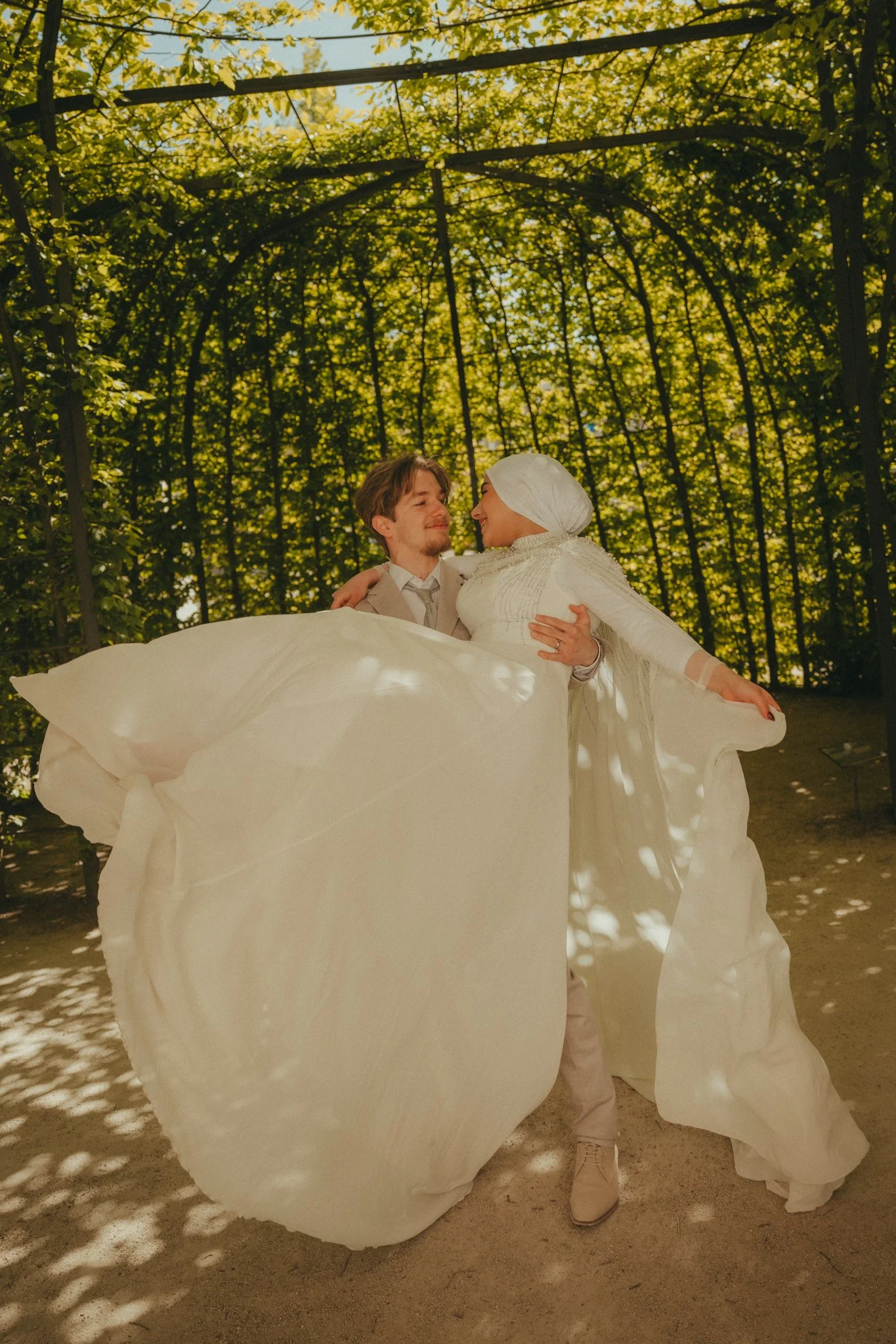 A couple dressed in white, with the woman in a modest wedding gown and headscarf and the man in a beige suit, dance outdoors in a lush green park, with sunlight filtering through trees.