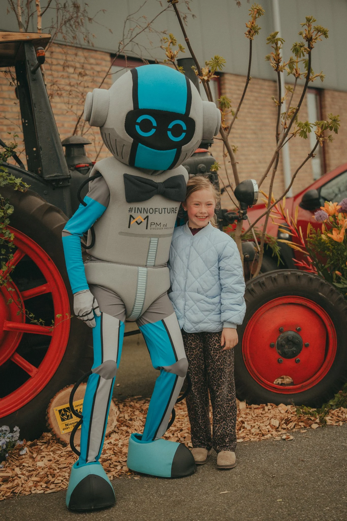A person in a robot costume standing next to a young girl outdoors in front of a tractor with red wheels and a brick building. The girl is smiling and wearing a light-colored quilted jacket and leopard print pants. There are small trees and leaves on