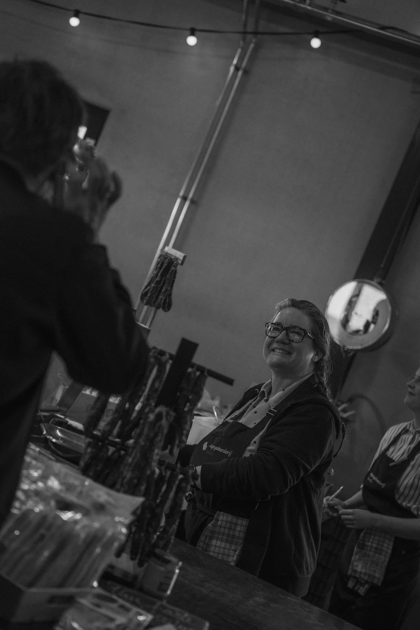 A woman smiling in a kitchen or café setting, wearing glasses and an apron, with people around her engaged in food preparation or serving, a mirror, string lights, and kitchen utensils visible in the background.