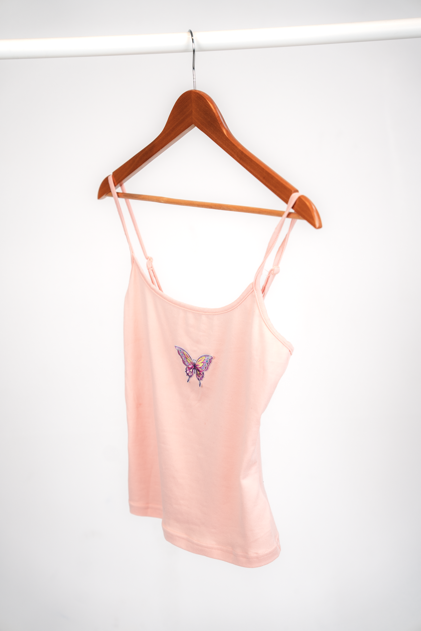 Pink sleeveless top with a butterfly embroidery on the front, hanging on a wooden hanger. The hanger is on a white clothing rod against a plain white background.