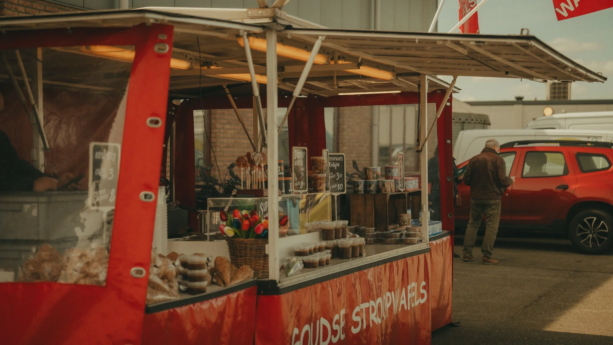 A food stand at an outdoor market with various food items and signs displaying prices, with a man and a parked red vehicle in the background.