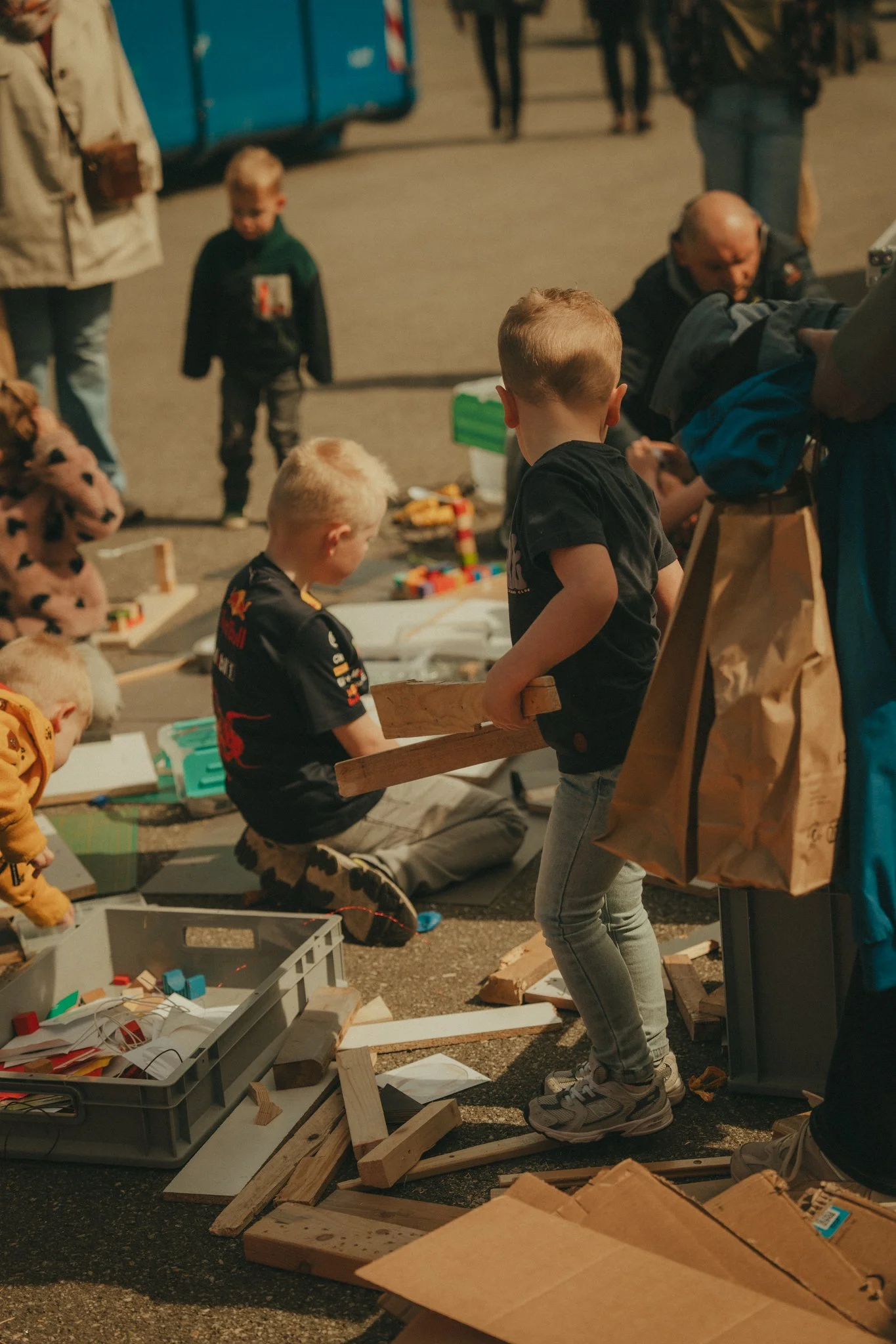 Children and adults participating in an outdoor building or craft activity surrounded by wooden pieces, tools, and containers.