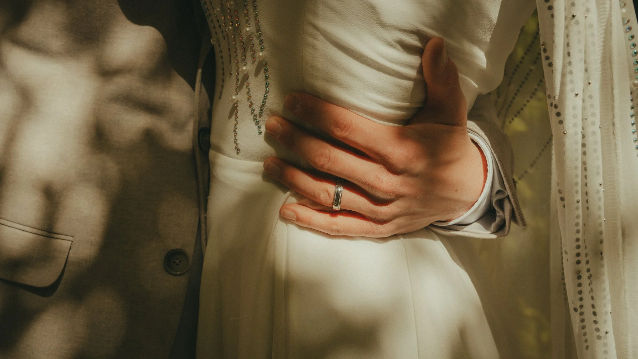 A close-up of a man's hand resting on a woman's waist, with the woman wearing a white outfit with embellishments, and a silver ring on the finger.