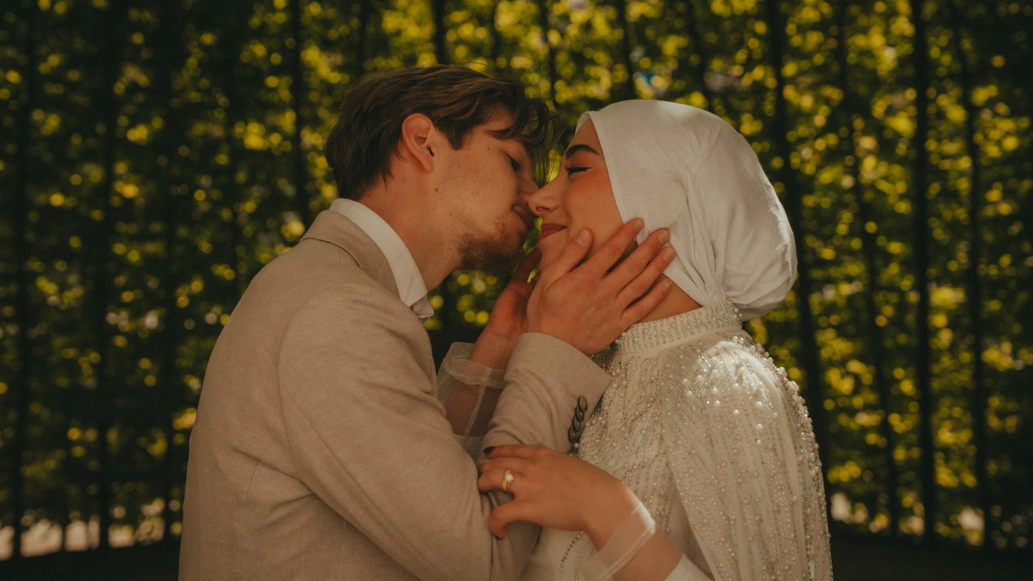 A couple sharing an intimate moment outdoors during sunset, facing each other with eyes closed, with trees in the background.