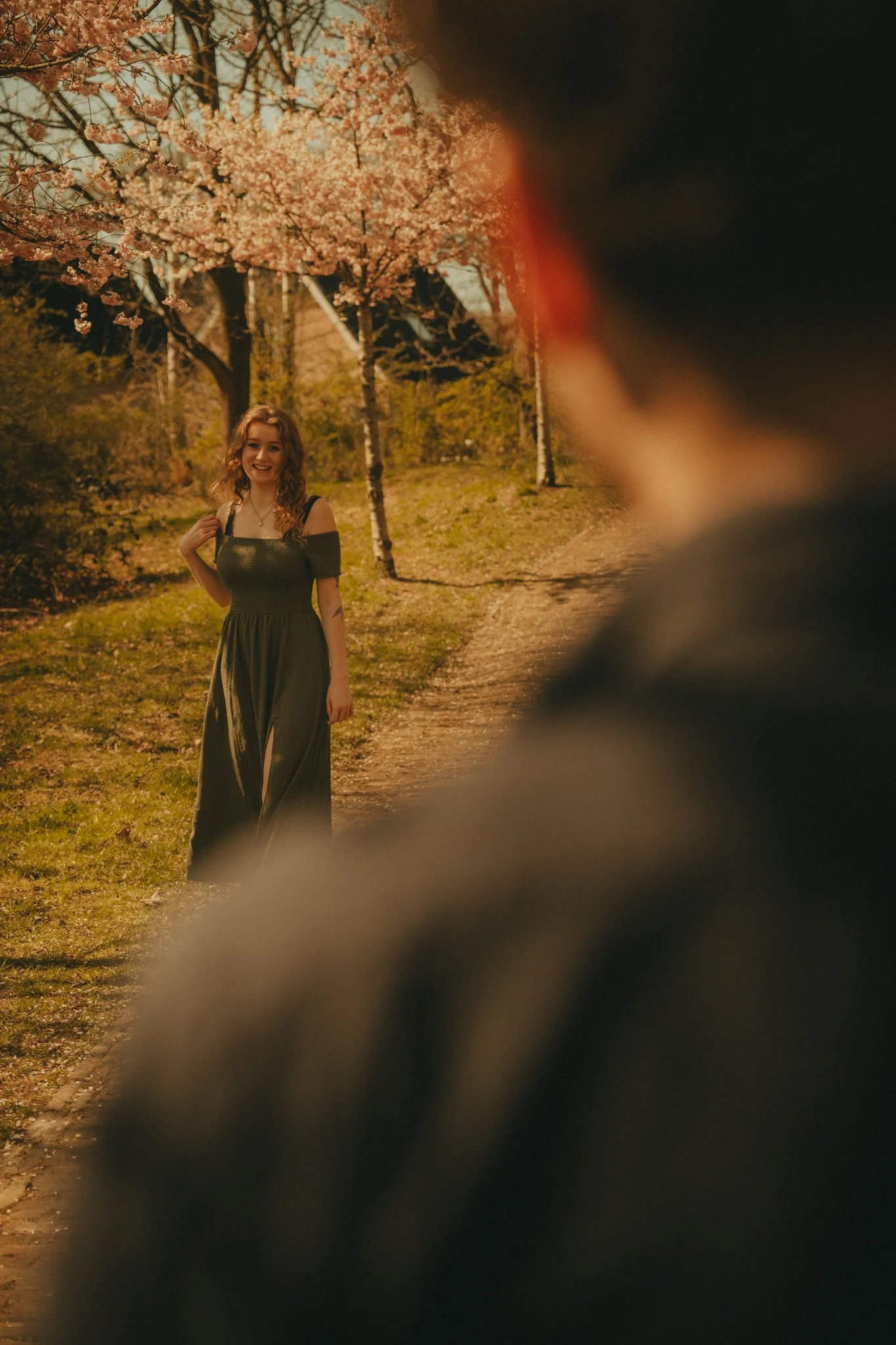 A woman with curly hair wearing a dark dress standing outdoors near blooming pink cherry blossom trees in daytime.