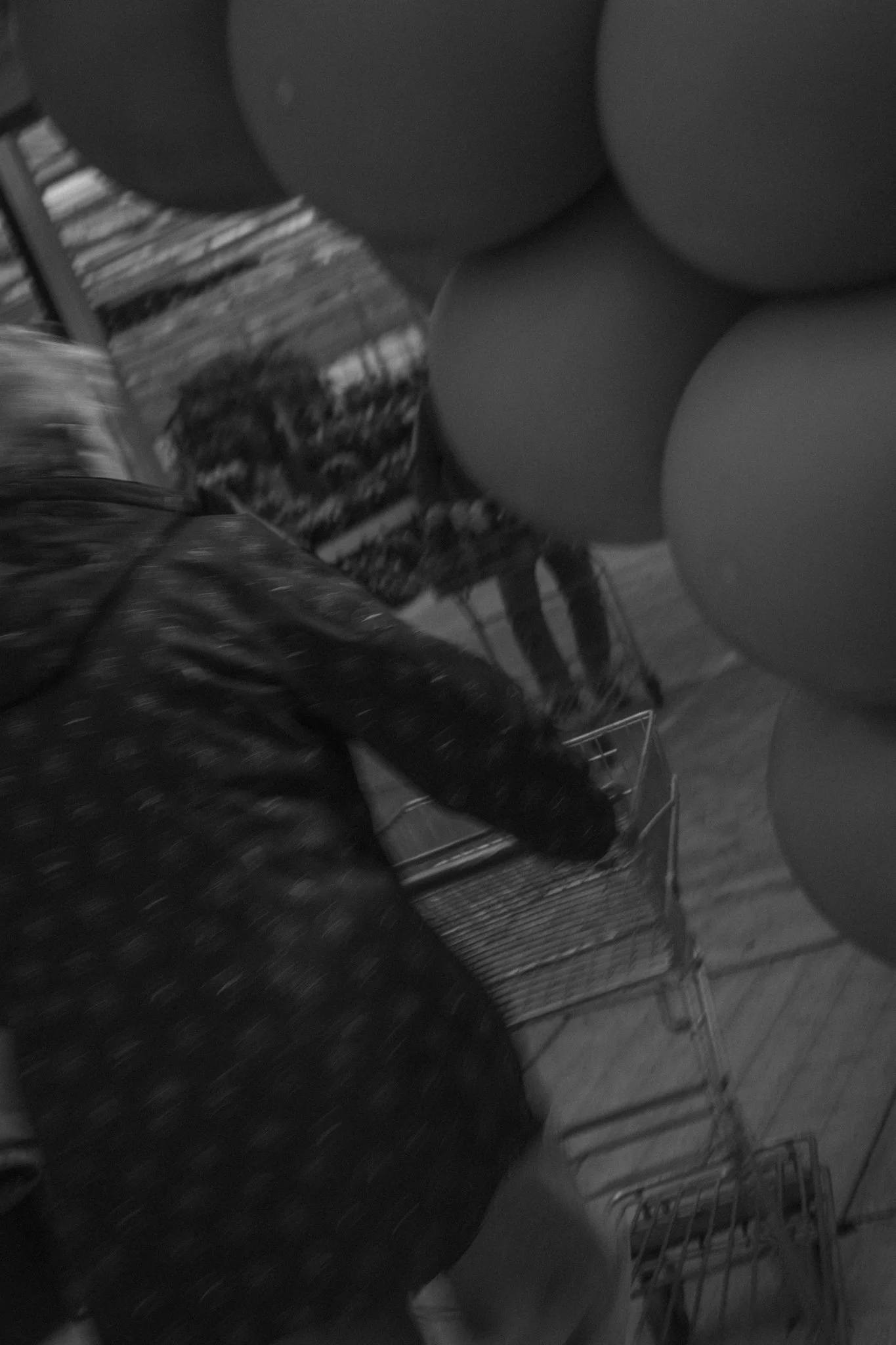 Black and white photo of a person with blonde hair, sitting at a table with a wire chair next to them, and several balloons in the background.