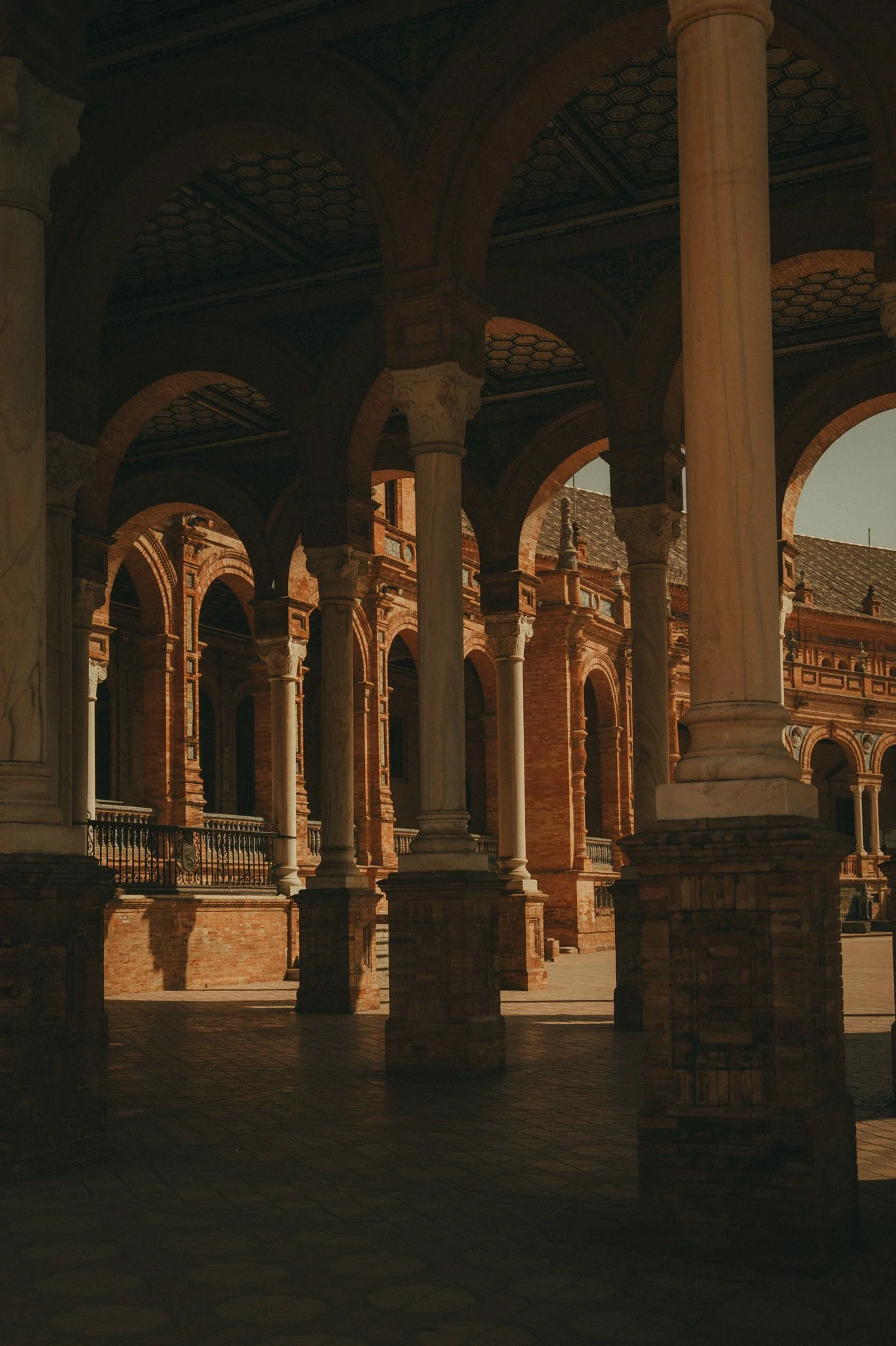 A historic architectural courtyard with stone columns supporting arches and a decorative ceiling, surrounded by brick walls with large windows.