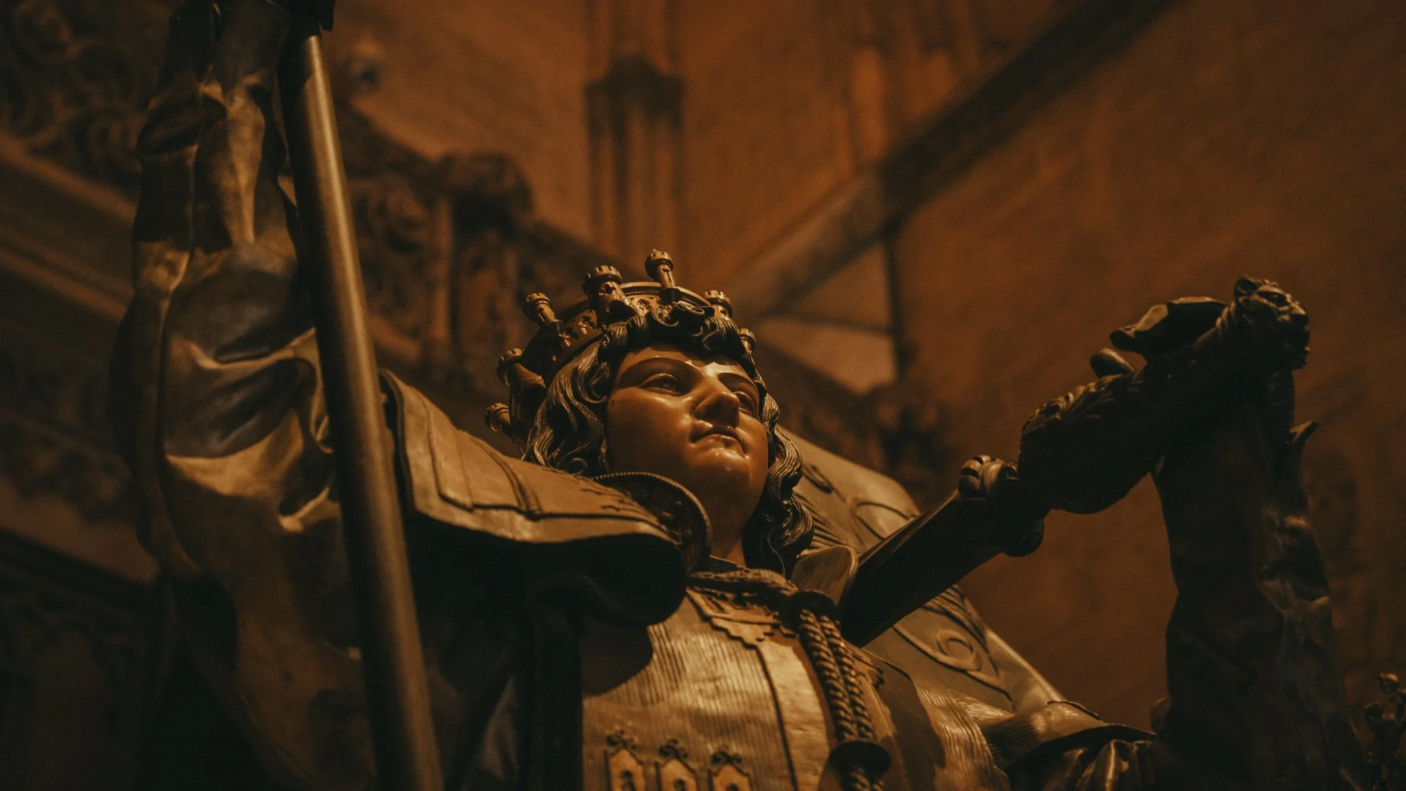 Close-up of a detailed bronze statue of a regal woman wearing a crown, with her arms outstretched inside a dimly lit room with ornate ceiling decorations.